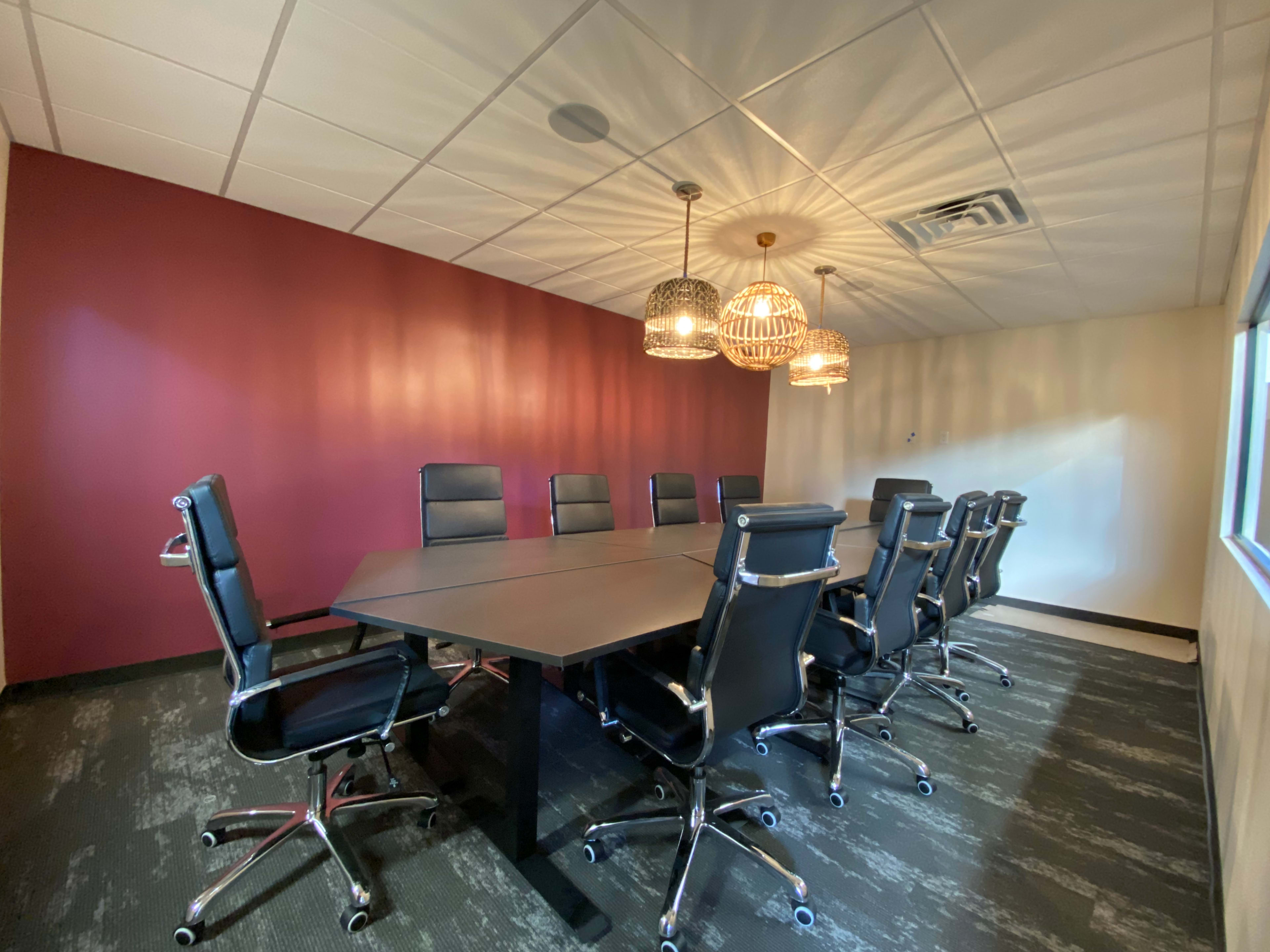 A conference room features a long, dark table surrounded by black office chairs and several pendant lights hanging from the ceiling.