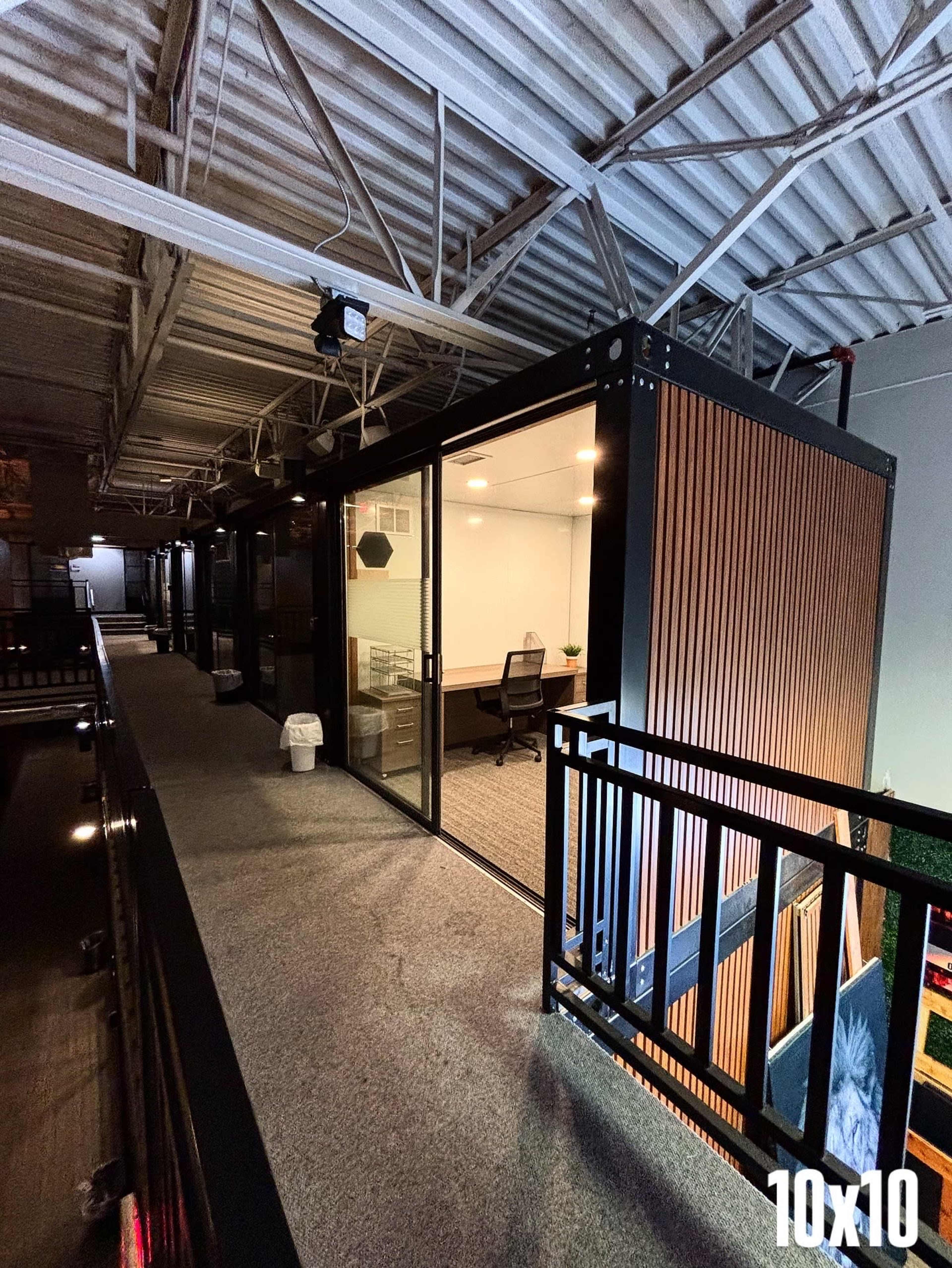 The image shows a modern office space within a shipping container, featuring large glass doors and wooden exterior panels, situated on an upper level of a building.