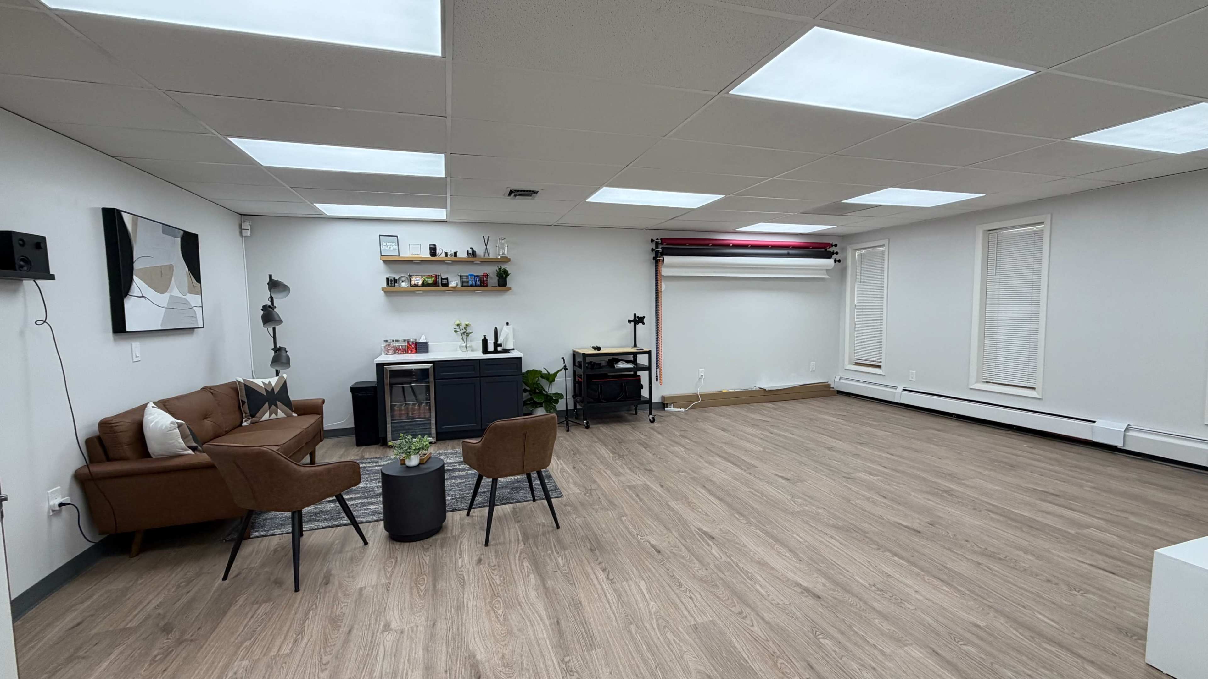 The image shows a spacious, well-lit room with a brown leather sofa, a small round table, and a dark cabinet against one wall, while a variety of windows and a backdrop system are visible on the opposite side.
