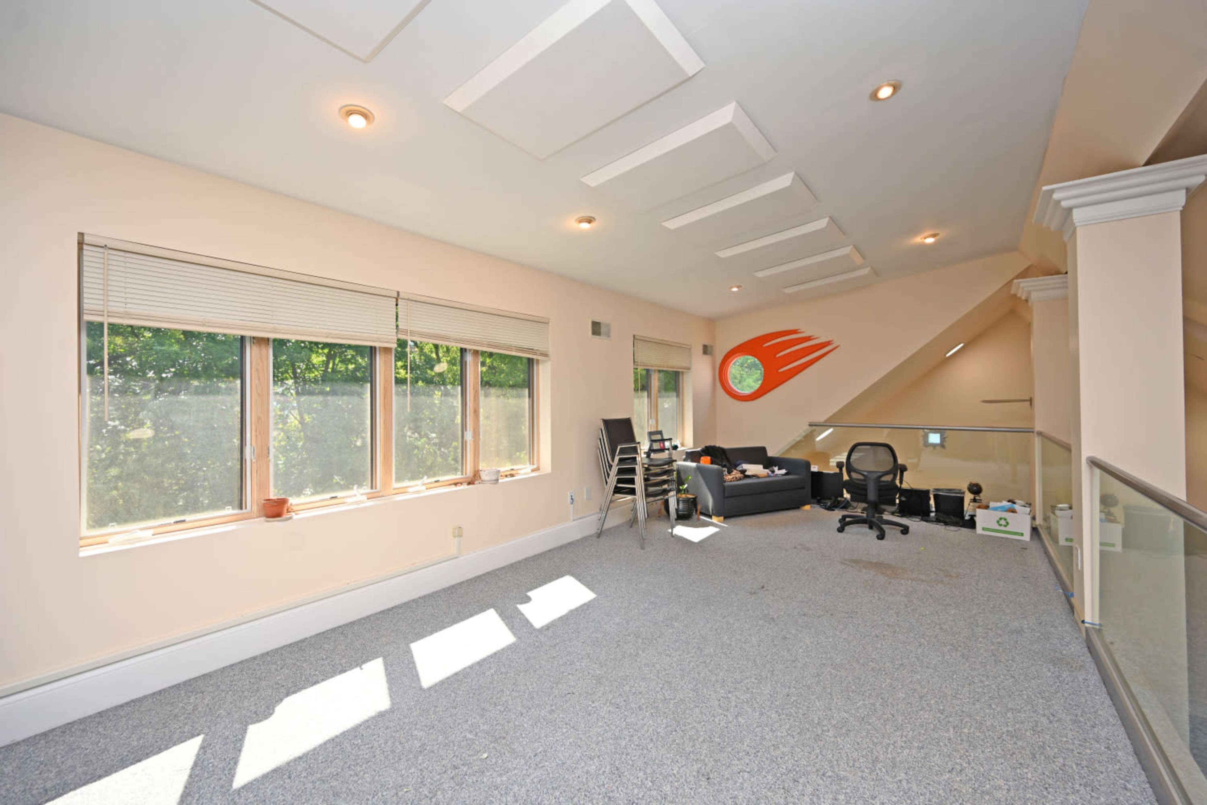 The image shows a spacious room with large windows, carpeted flooring, and minimal furniture including a chair and a couch.