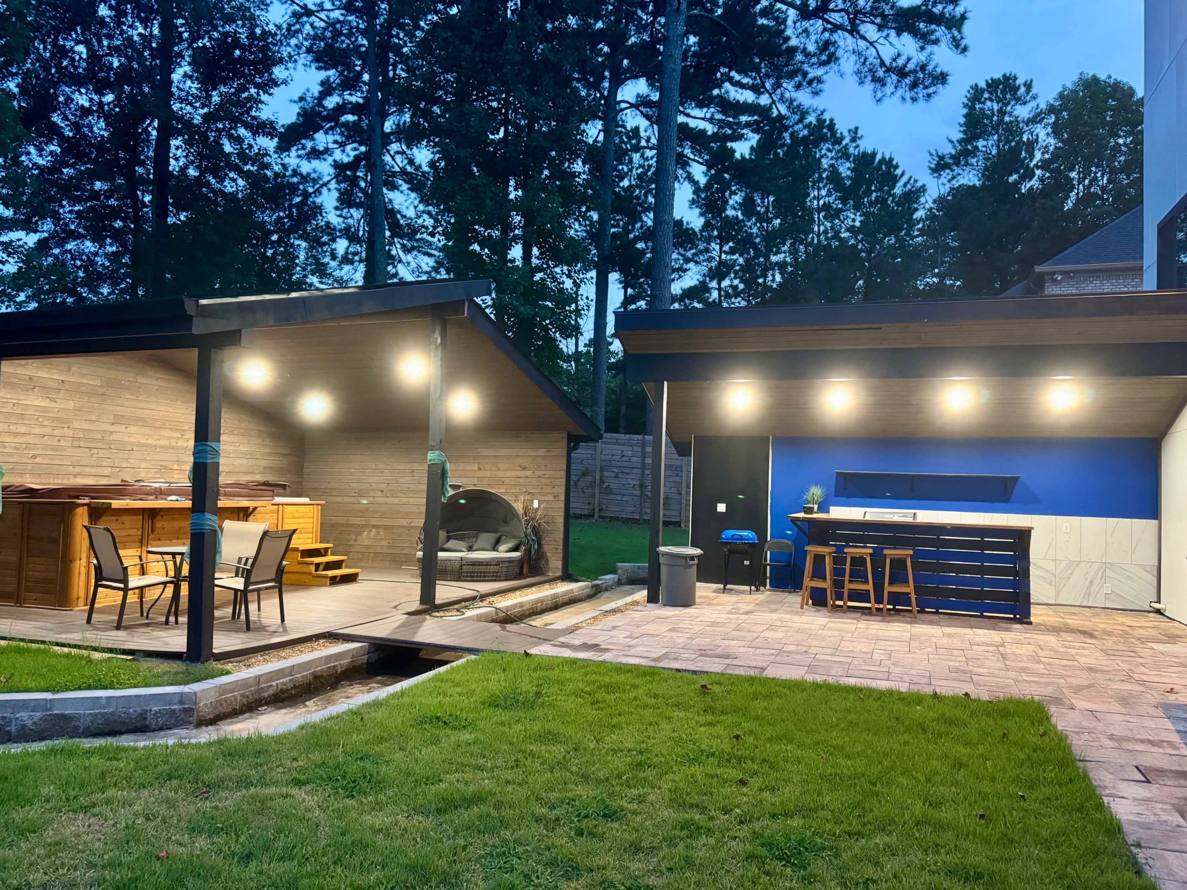The image shows a backyard with two structures: a gazebo on the left with chairs and a hot tub, and an outdoor kitchen on the right with a bar counter and a grill, all set on a grassy area.
