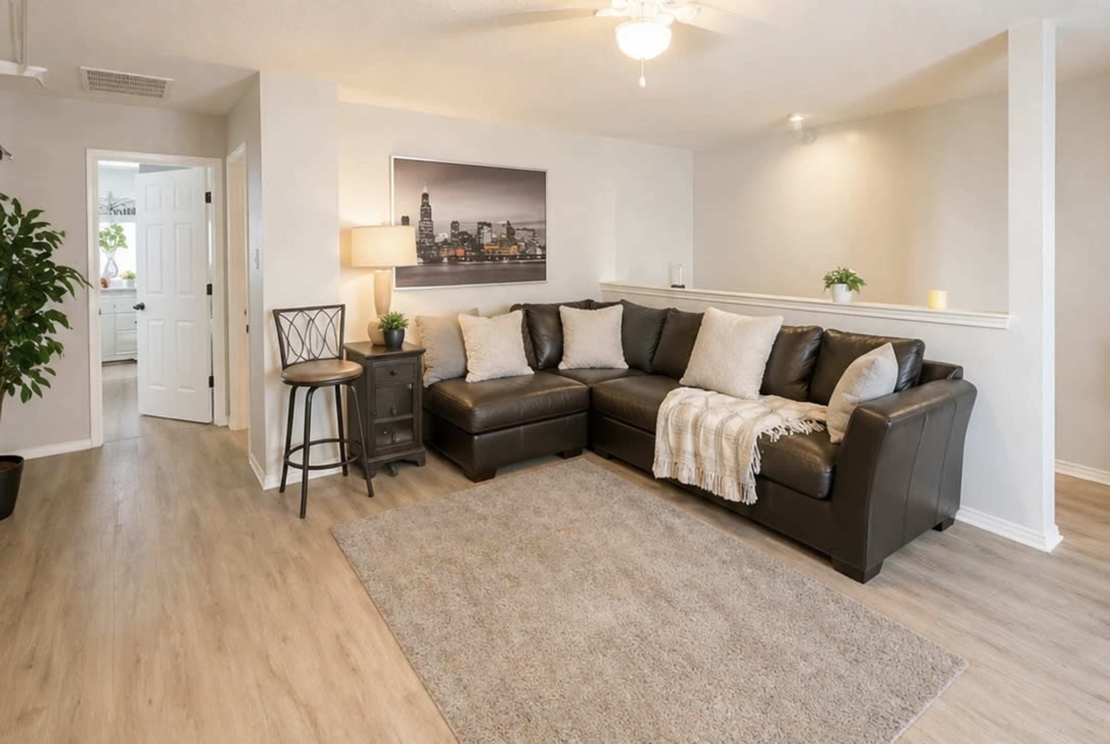 The image shows a modern living room featuring a dark leather sectional sofa, a small side table with a lamp, and light-colored walls with a rug on the floor.