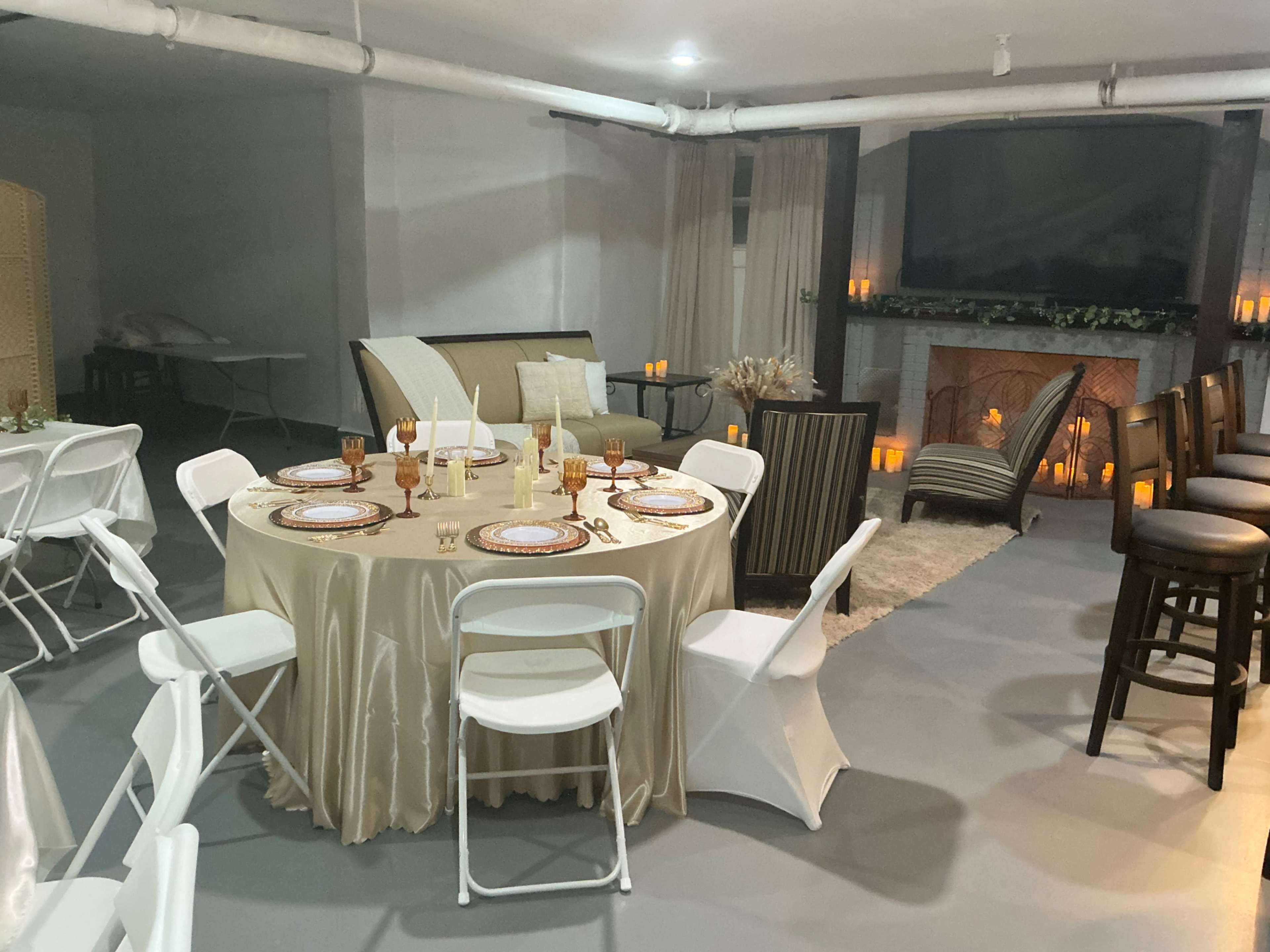 The image shows a decorated indoor space with a round table set for dinner, surrounded by white chairs, a lounge area with a sofa, and a fireplace adorned with candles.