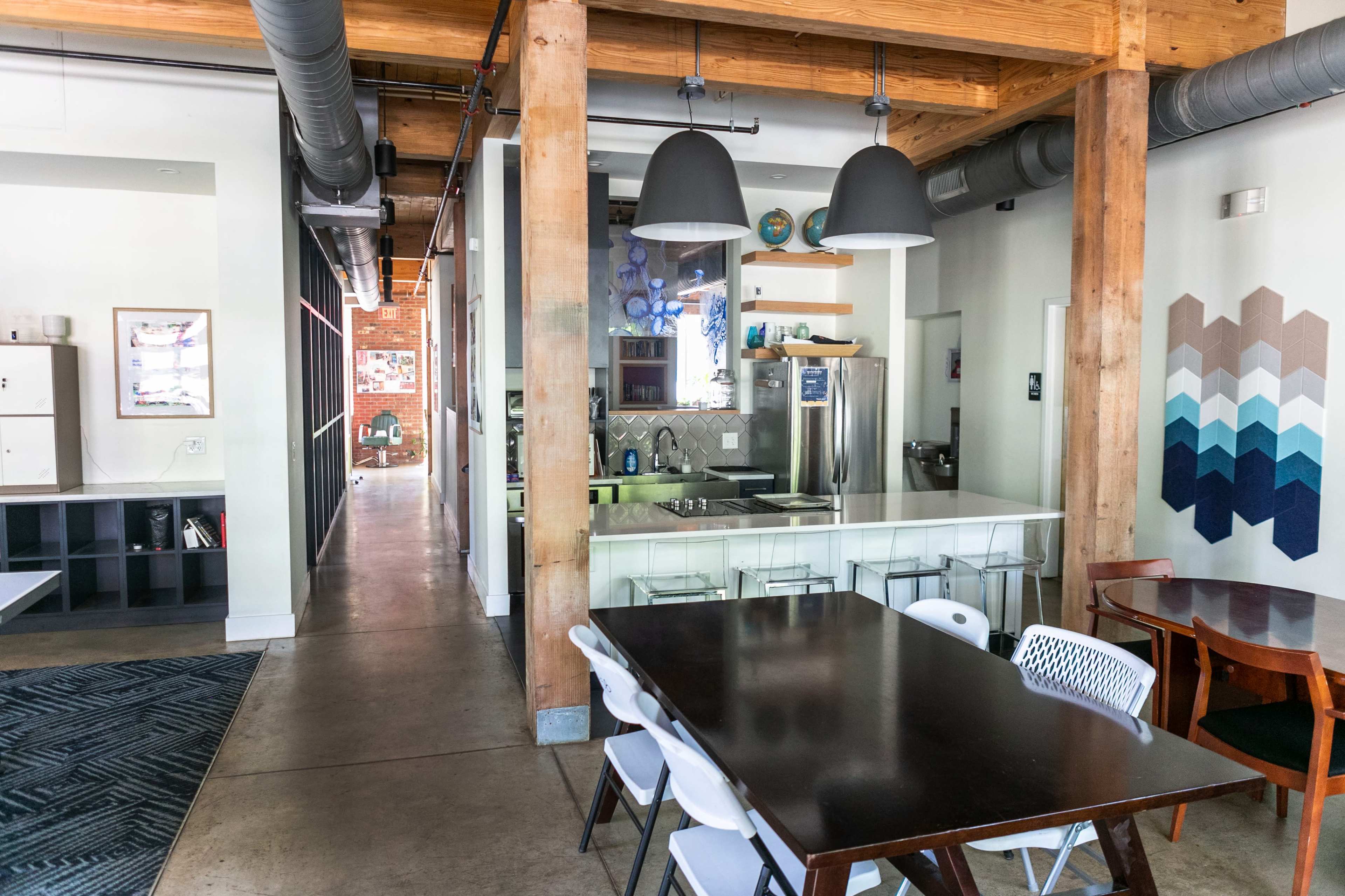 The image shows an open-concept space with wooden beams, a dining area featuring dark wooden tables and white chairs, and a kitchen area visible in the background.