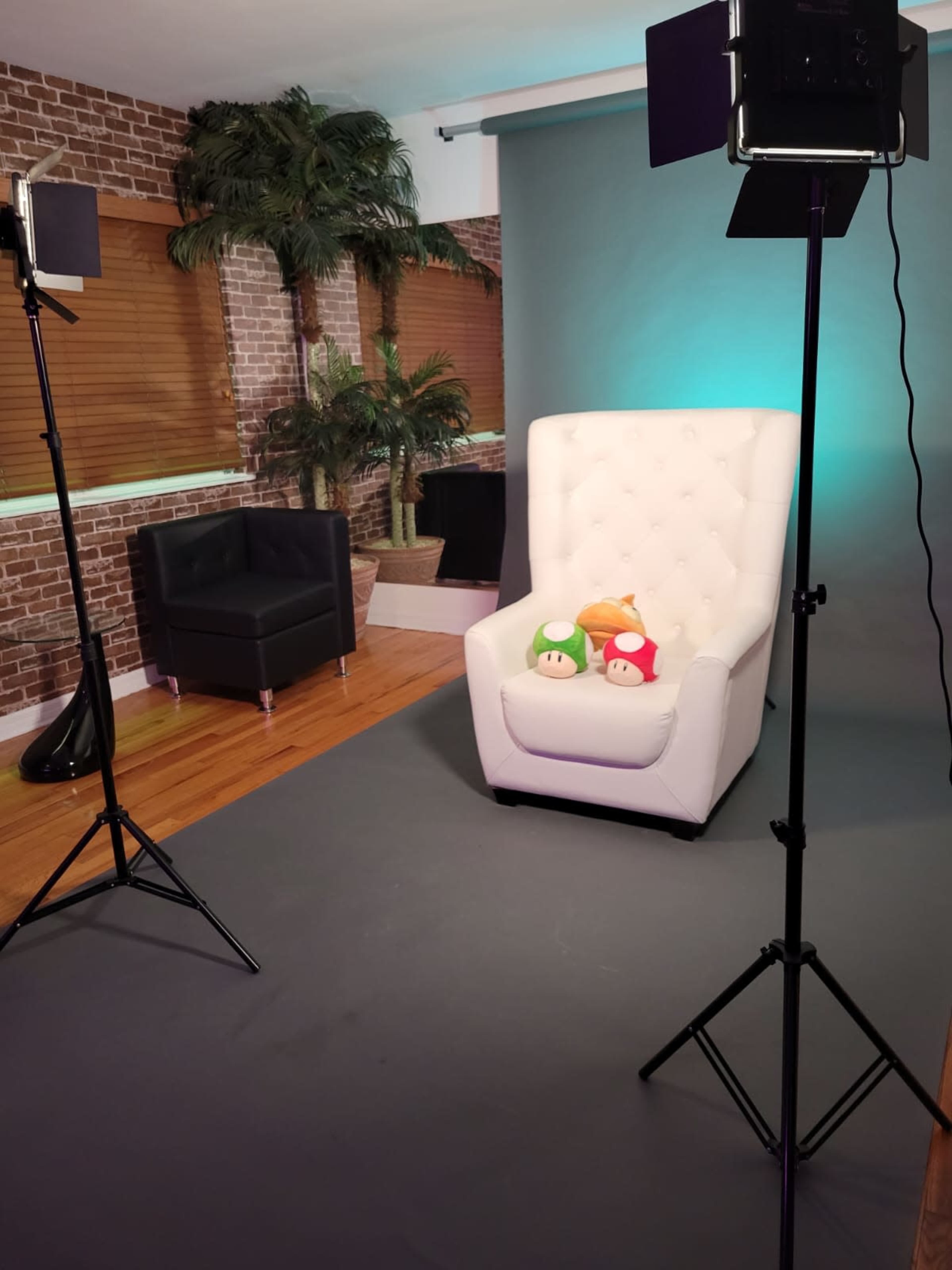 A brightly lit studio setting featuring a white tufted armchair with three colorful plush mushroom toys seated on it, surrounded by potted plants and studio lighting equipment.