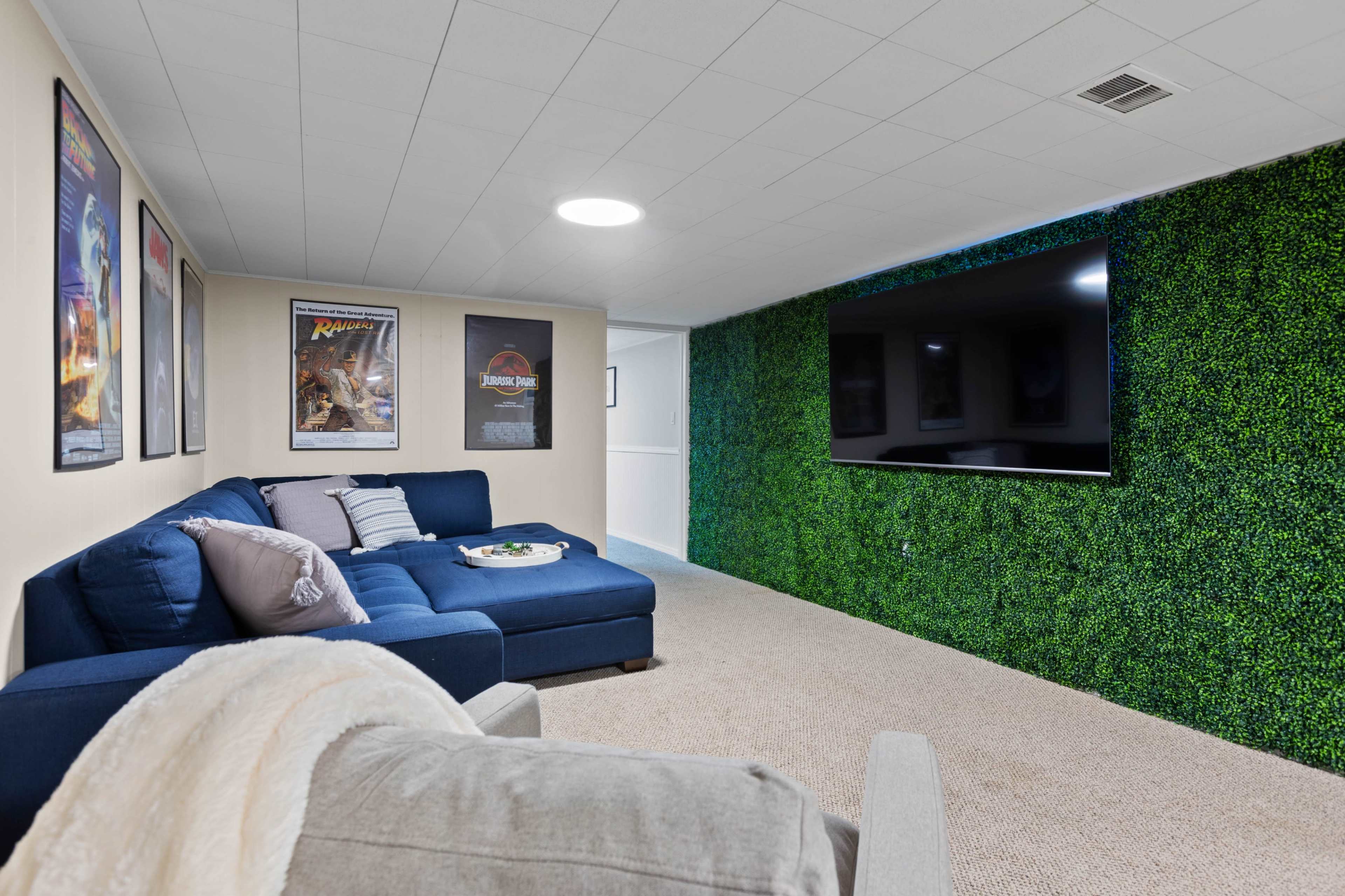 A basement recreation area featuring a blue sectional sofa, a large wall-mounted TV, and a wall adorned with green faux foliage, along with framed movie posters on the opposite wall.