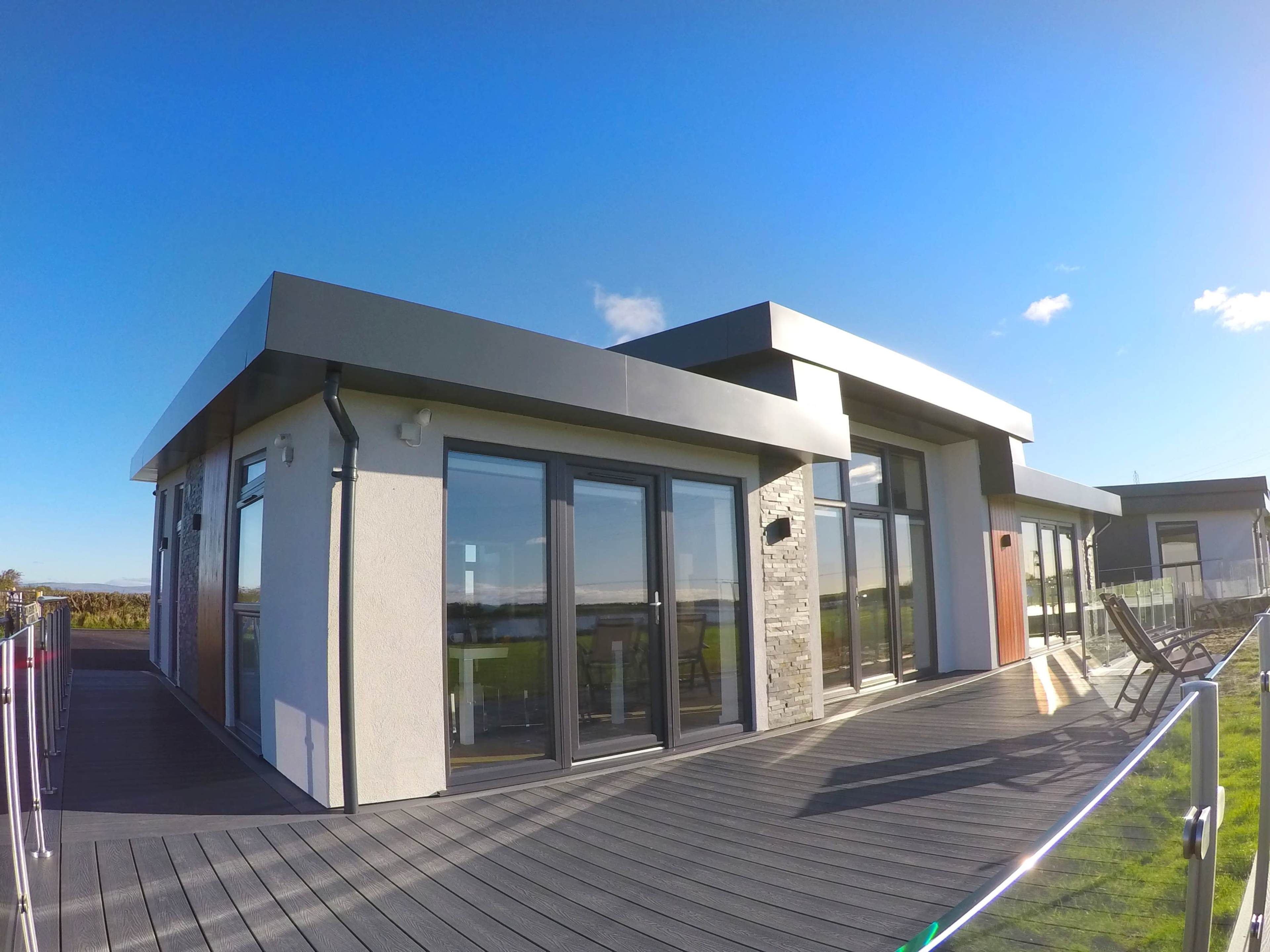 A modern house with large windows and a flat roof is situated on a balcony overlooking a grassy landscape under a clear blue sky.