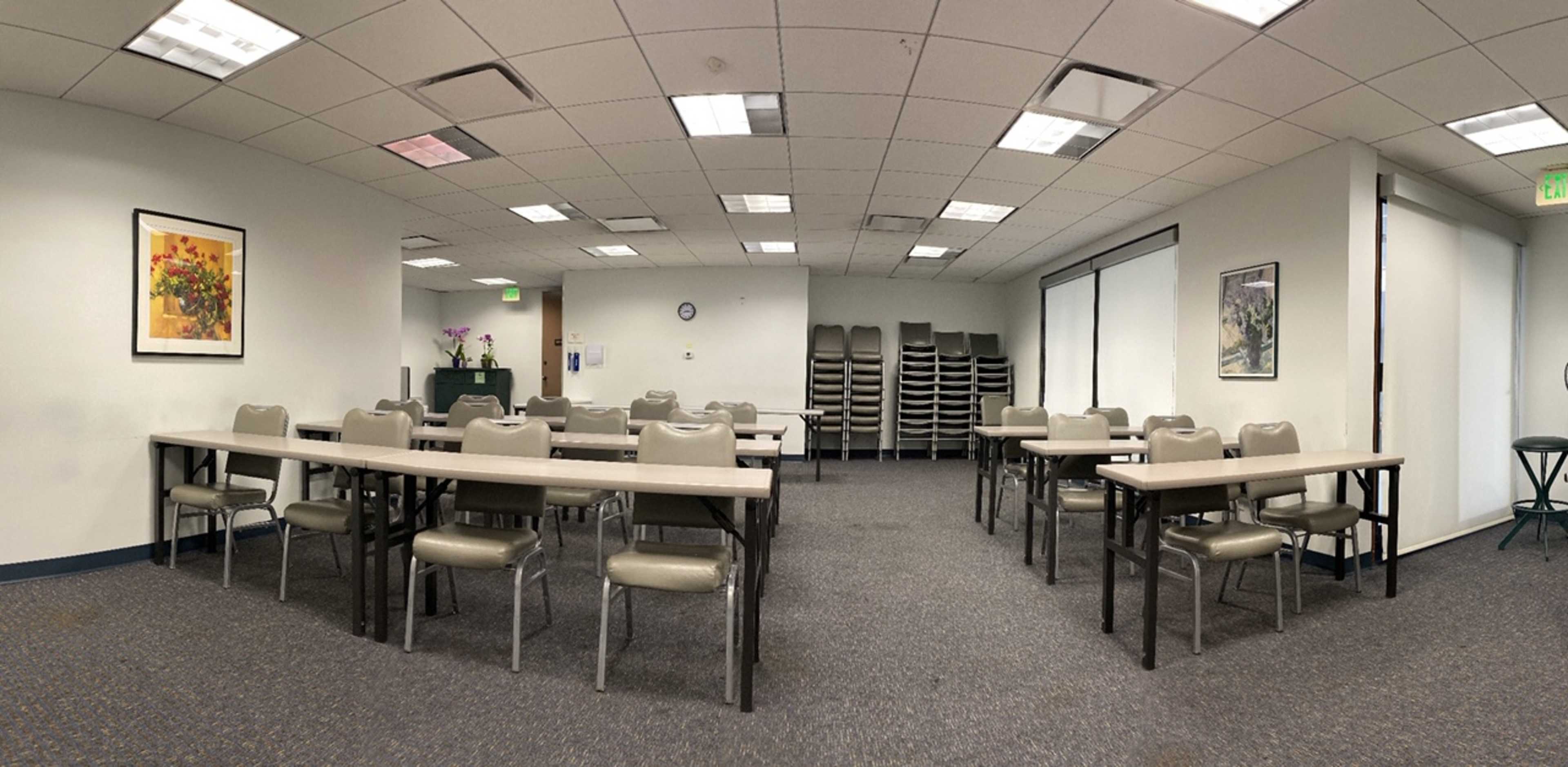 A sparsely furnished meeting room with several tables and chairs arranged neatly, and a stack of chairs against the back wall.