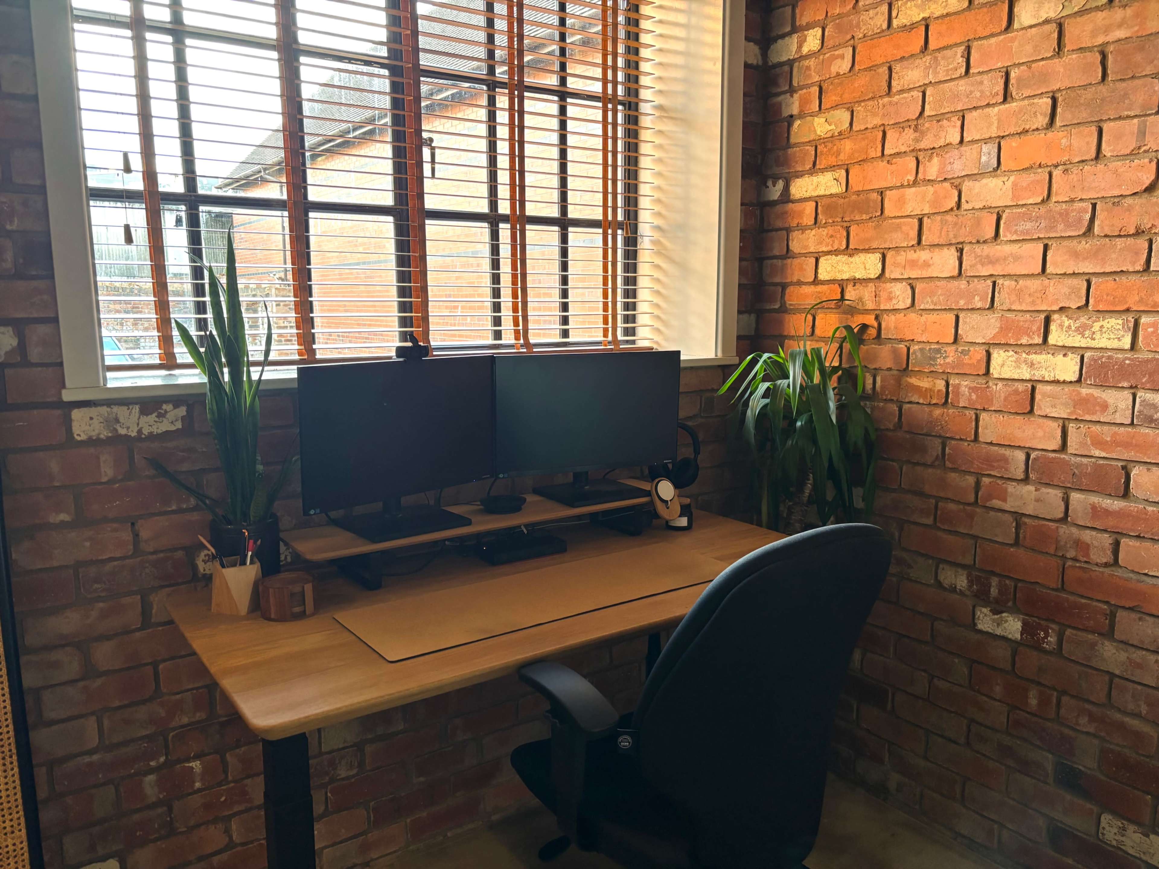 The image shows a home office setup with a wooden desk, two computer monitors, a black ergonomic chair, and potted plants against a brick wall.