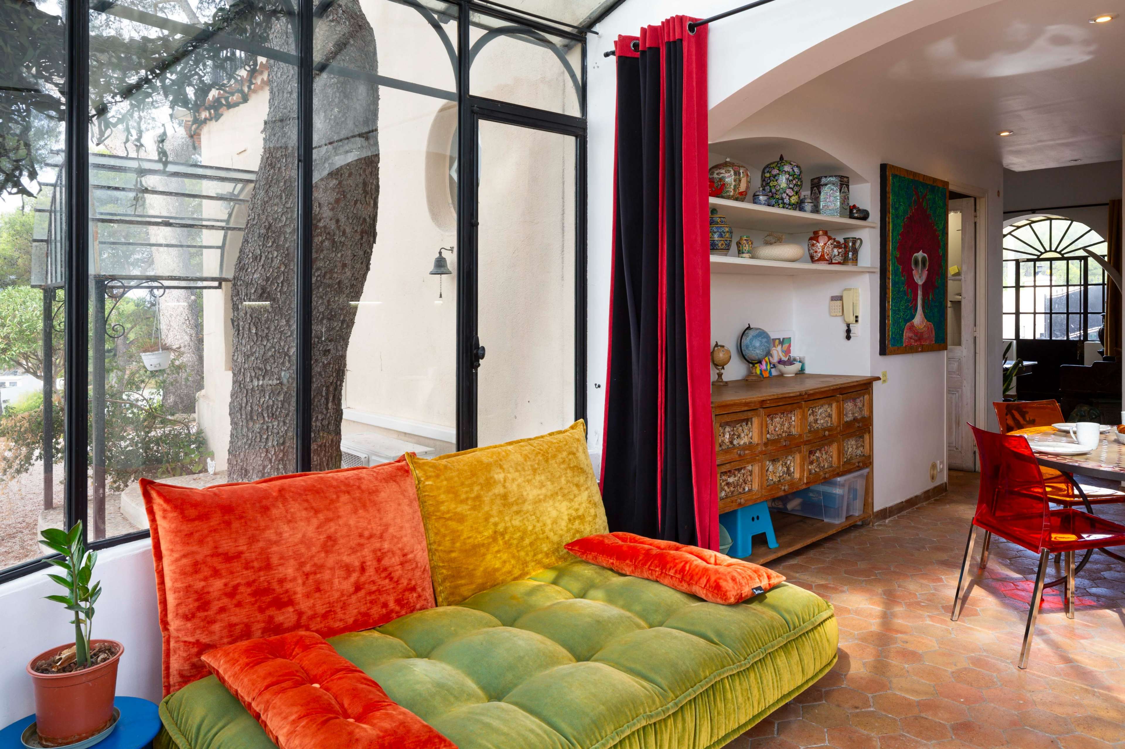 The image depicts a cozy interior space with a colorful sofa, a decorative wooden sideboard, and large windows that offer a view of the outdoors.