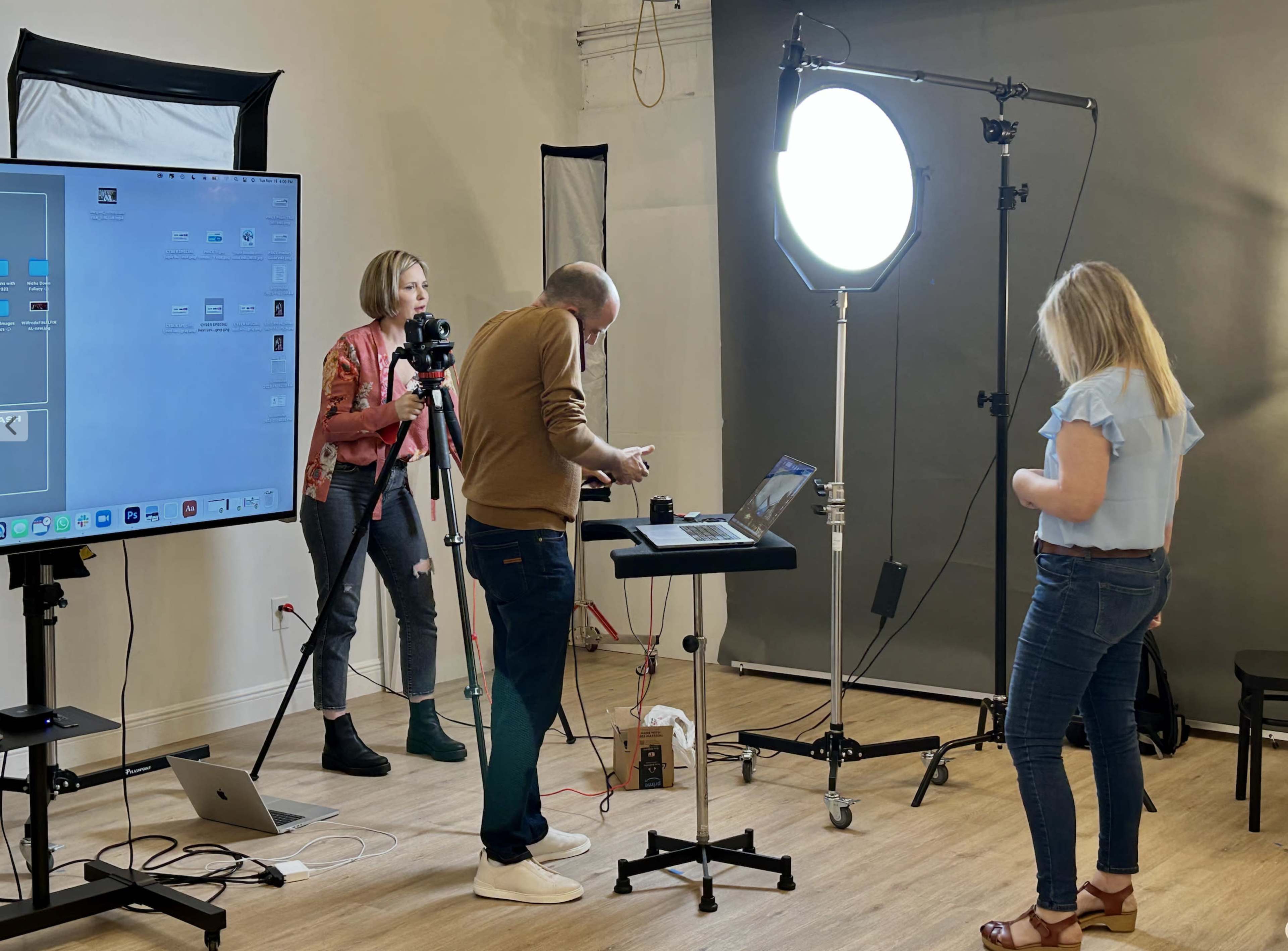 A small studio setting features a man adjusting a laptop on a table while a woman prepares for a shot with a camera, and another woman stands nearby near lighting equipment.