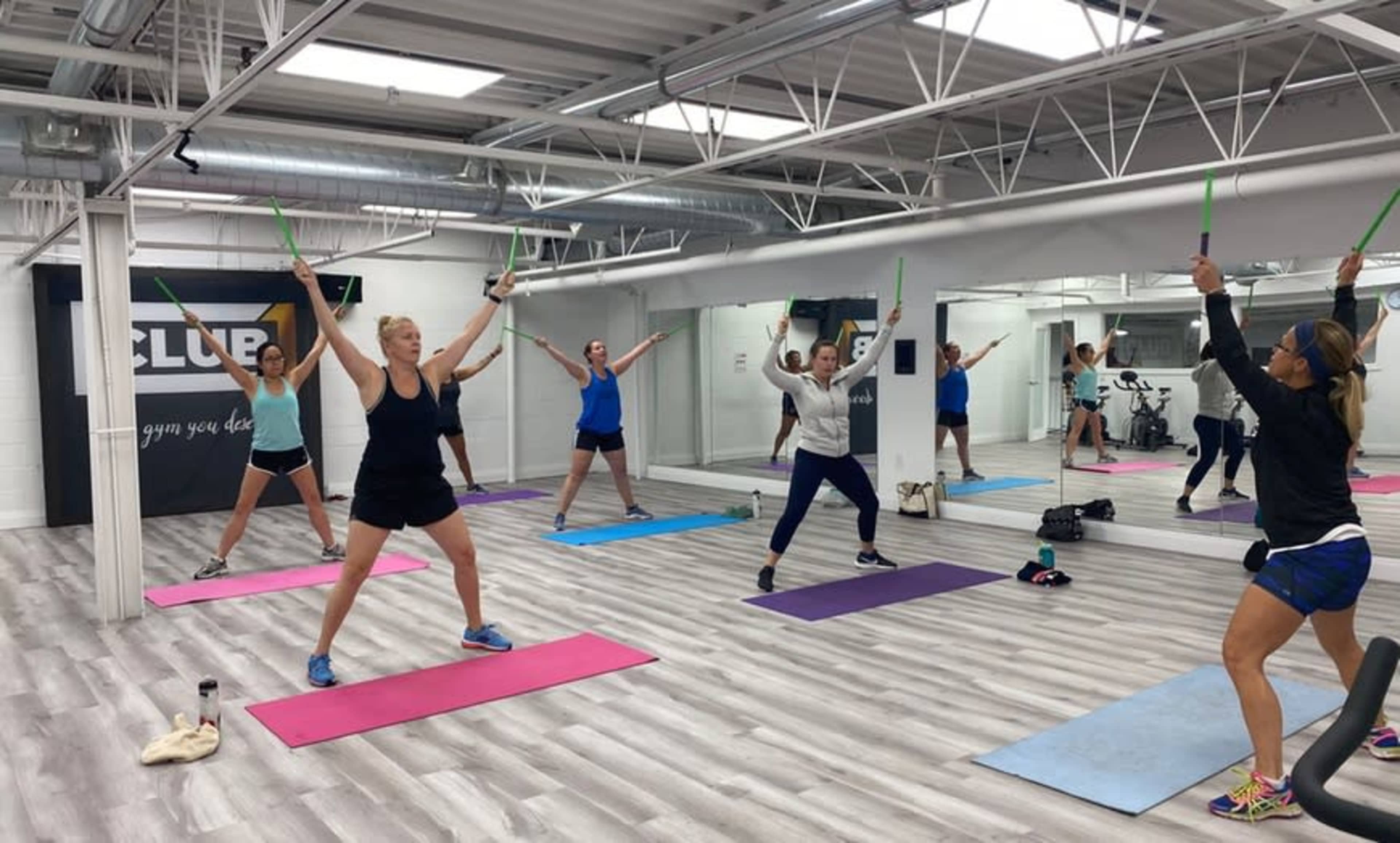 A group of individuals are participating in a fitness class, utilizing sticks while performing exercises on yoga mats in a mirrored studio.