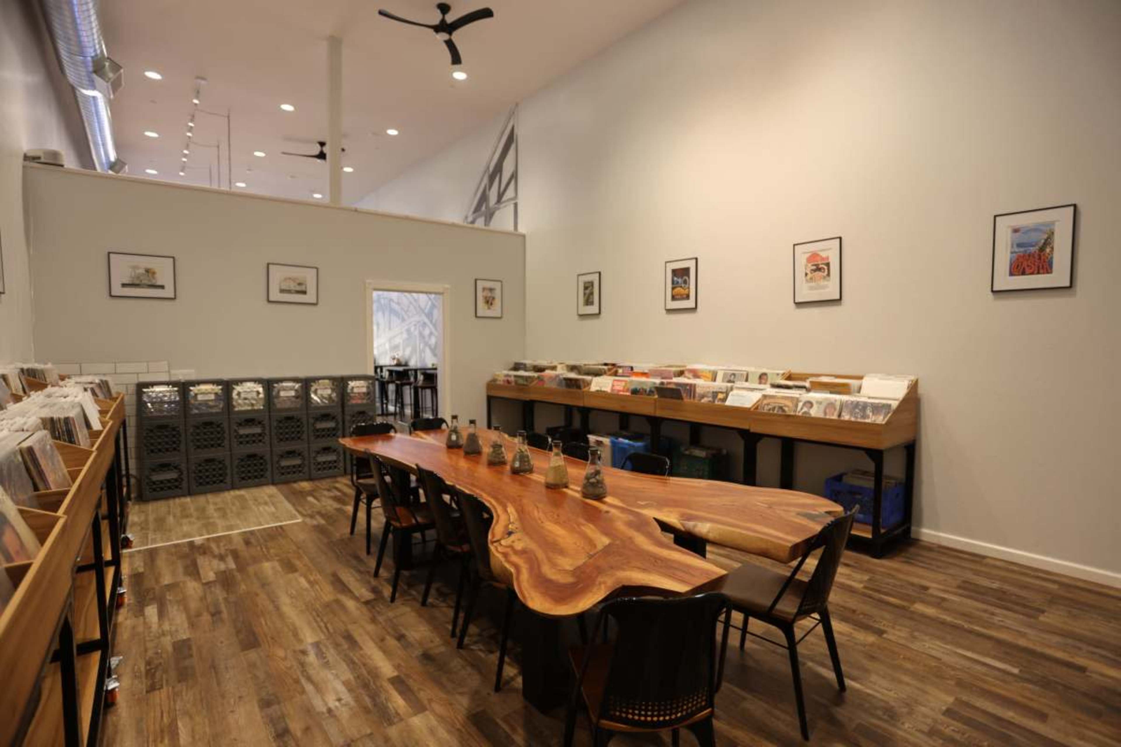 The image shows a spacious interior of a modern shop with a long wooden table, several chairs, and shelves of various items along the walls.