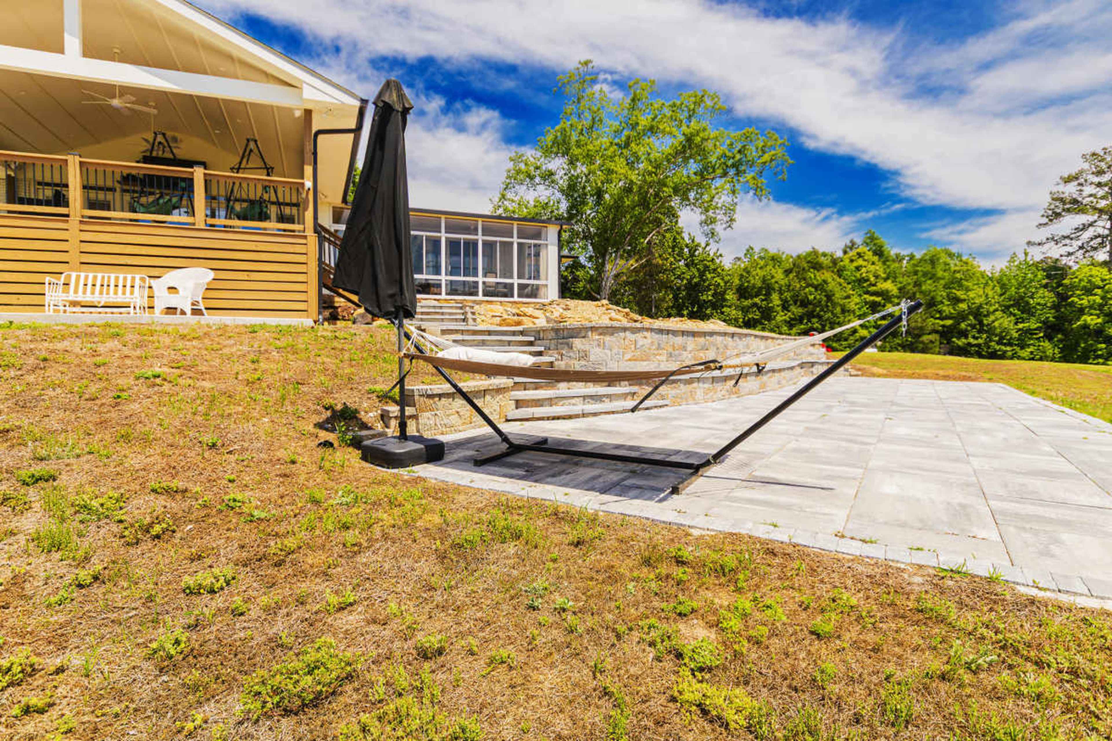 A hammock stand is set up on a stone patio beside a wooden house with a porch, surrounded by grassy land and trees under a cloudy sky.