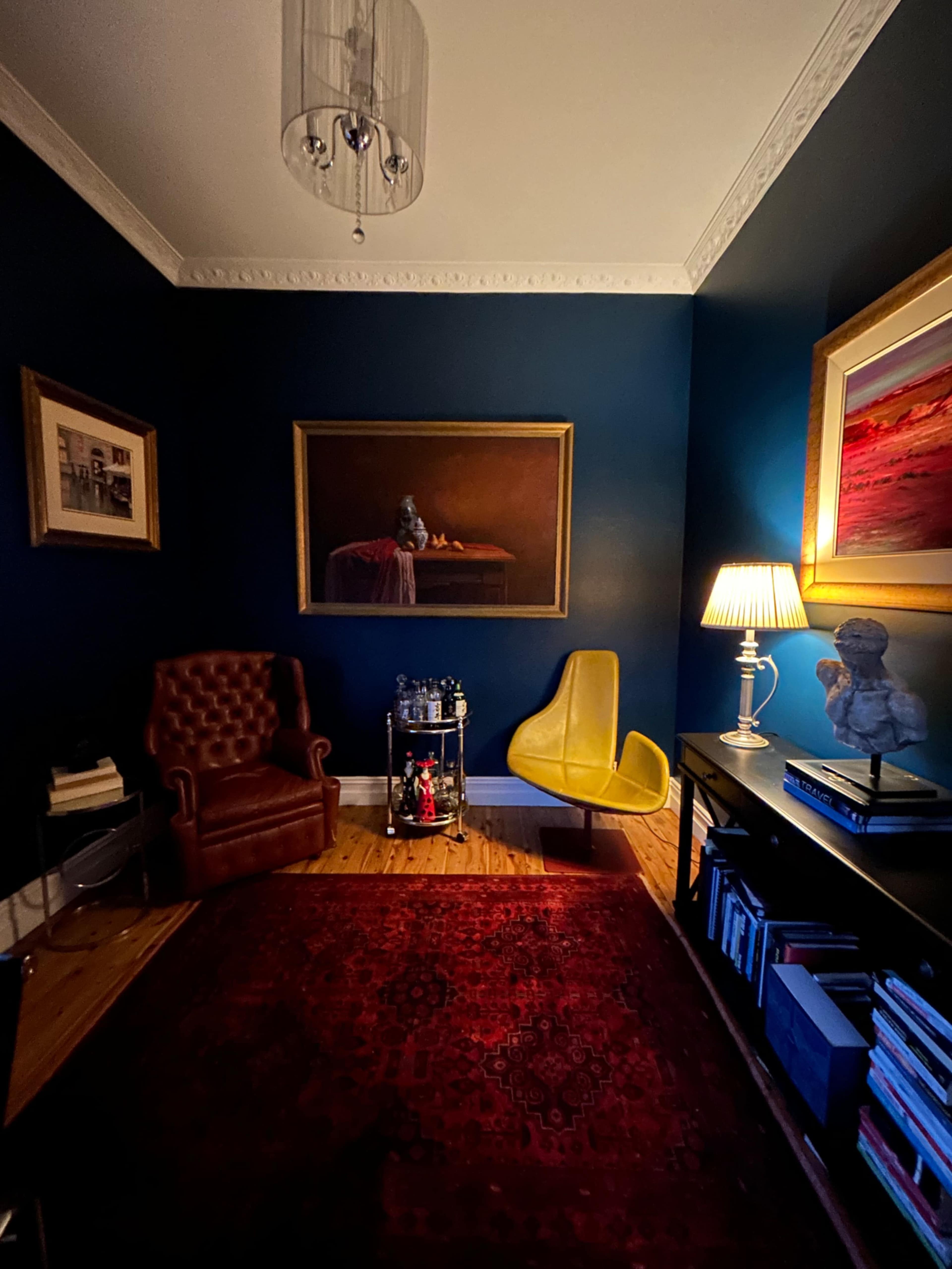 The room features a dark blue wall, a leather armchair, a yellow chair, a lamp, and various artworks, with a rug covering the wooden floor.