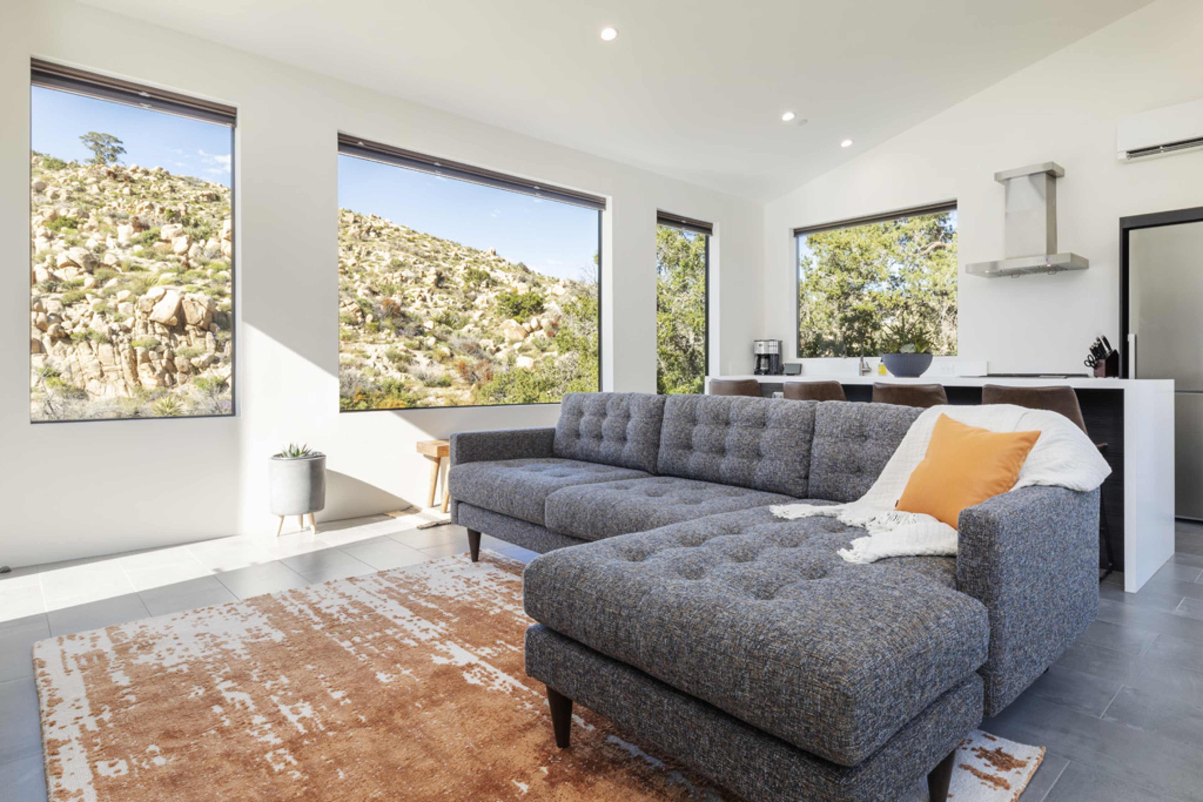 A spacious living room features a gray sectional couch, large windows with views of a rocky landscape, and minimalistic decor.