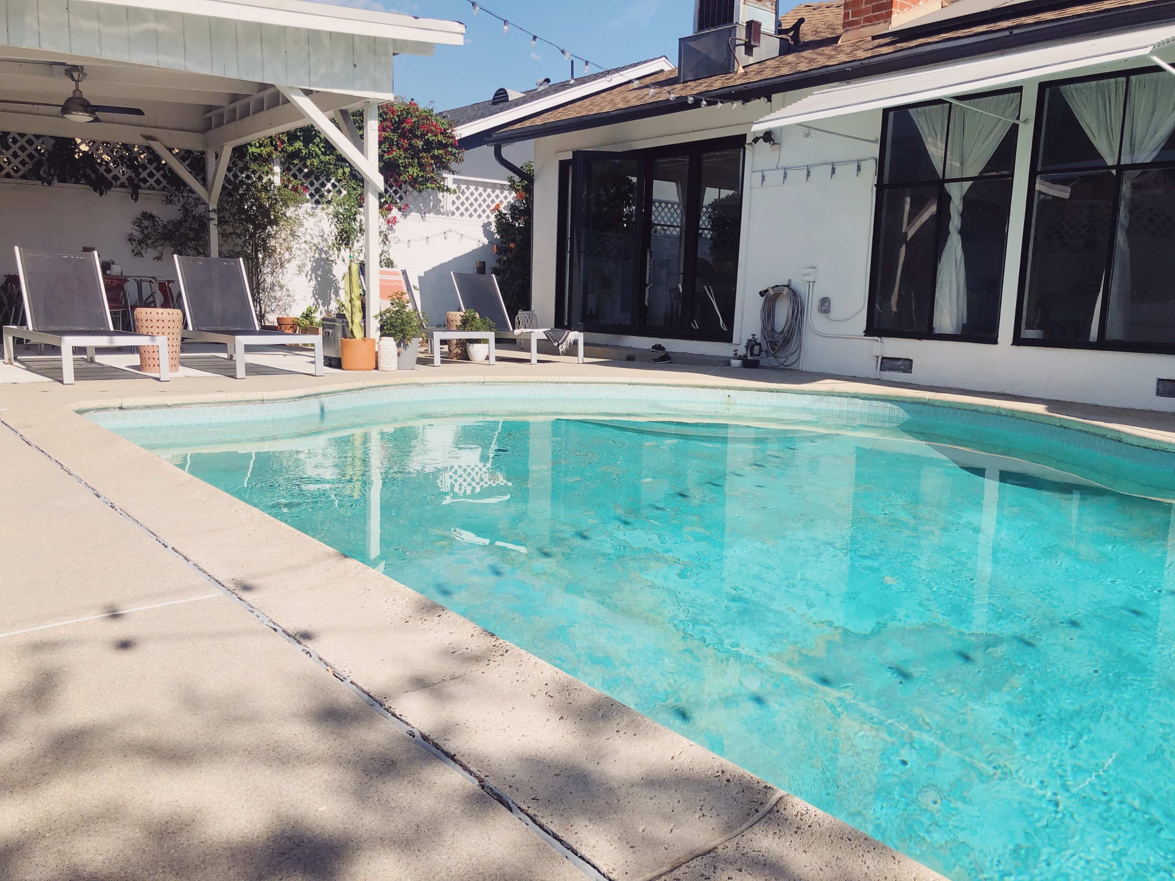 A clear swimming pool is surrounded by lounge chairs and a house with large windows under a sunny sky.