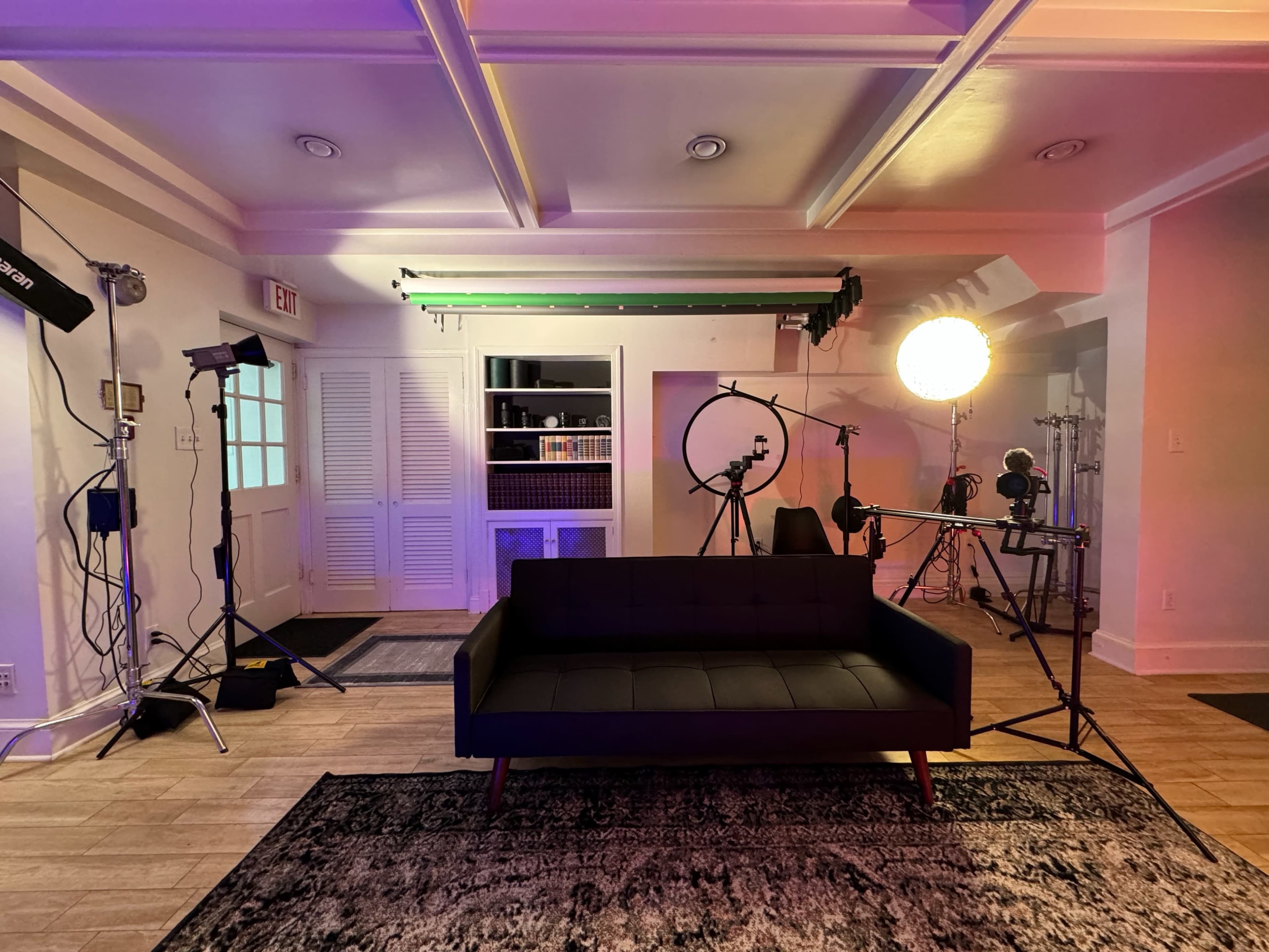 The image shows a modern studio setup featuring a black sofa on a rug, with various lighting equipment and a backdrop positioned in the background.