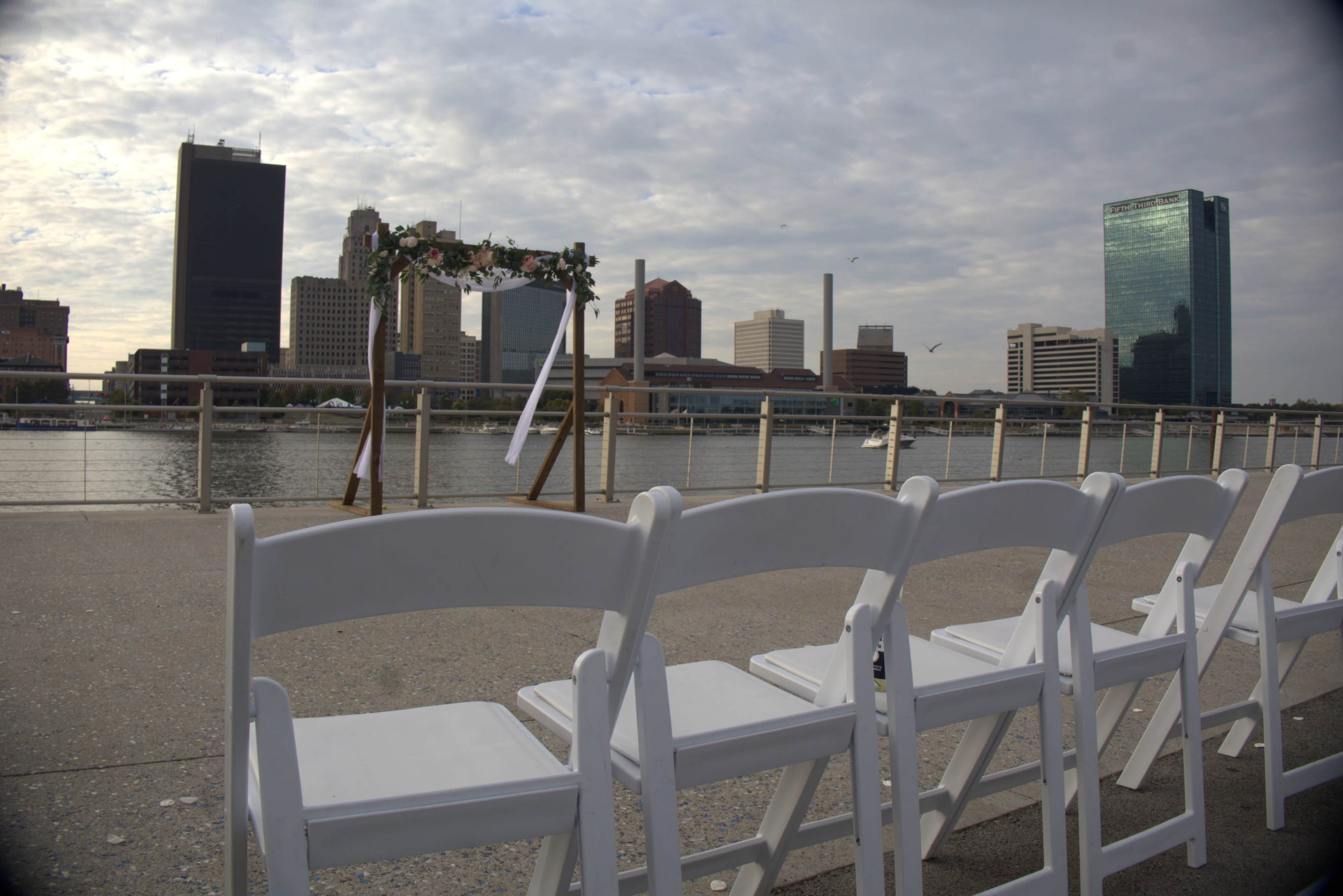 The scene shows empty white chairs set up along a waterfront with a decorative arch in the background, overlooking a city skyline under a cloudy sky.