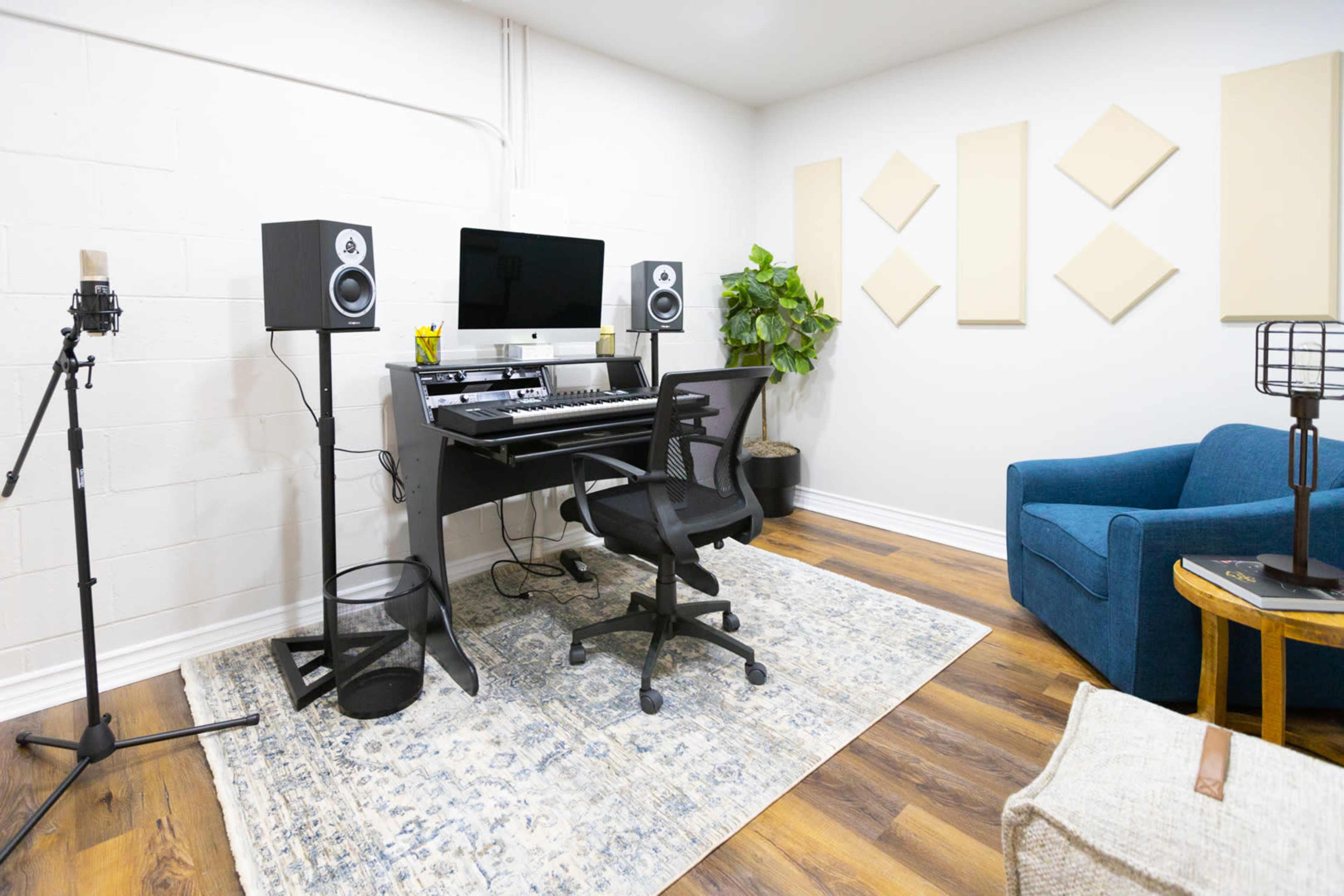 The image shows a modern music studio with a computer setup, a chair, a microphone stand, plants, and soft furnishings against a light-colored wall.