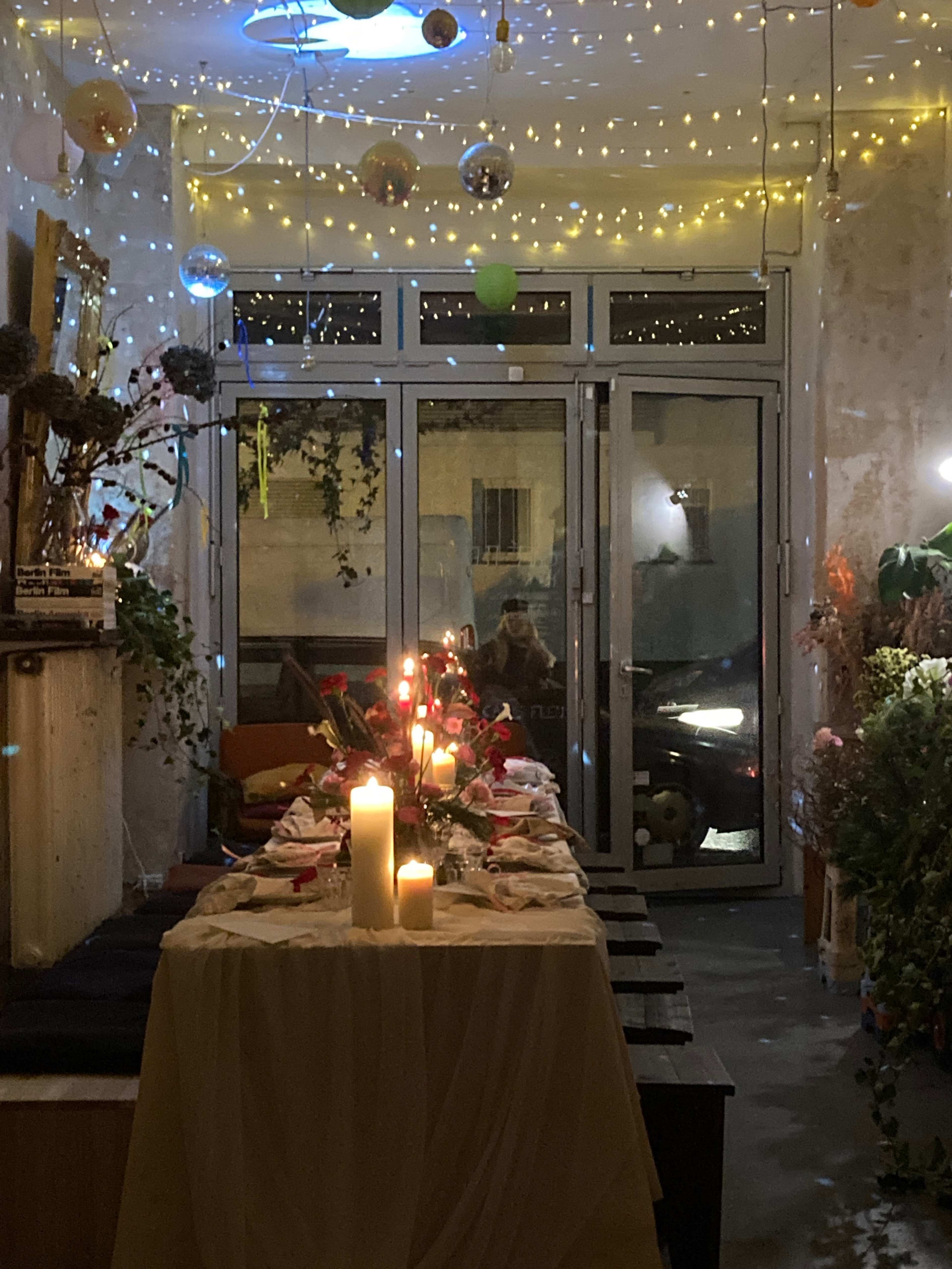 A long table is set for a dinner, adorned with candles and flowers, in a dimly lit space decorated with string lights and colorful ornaments.