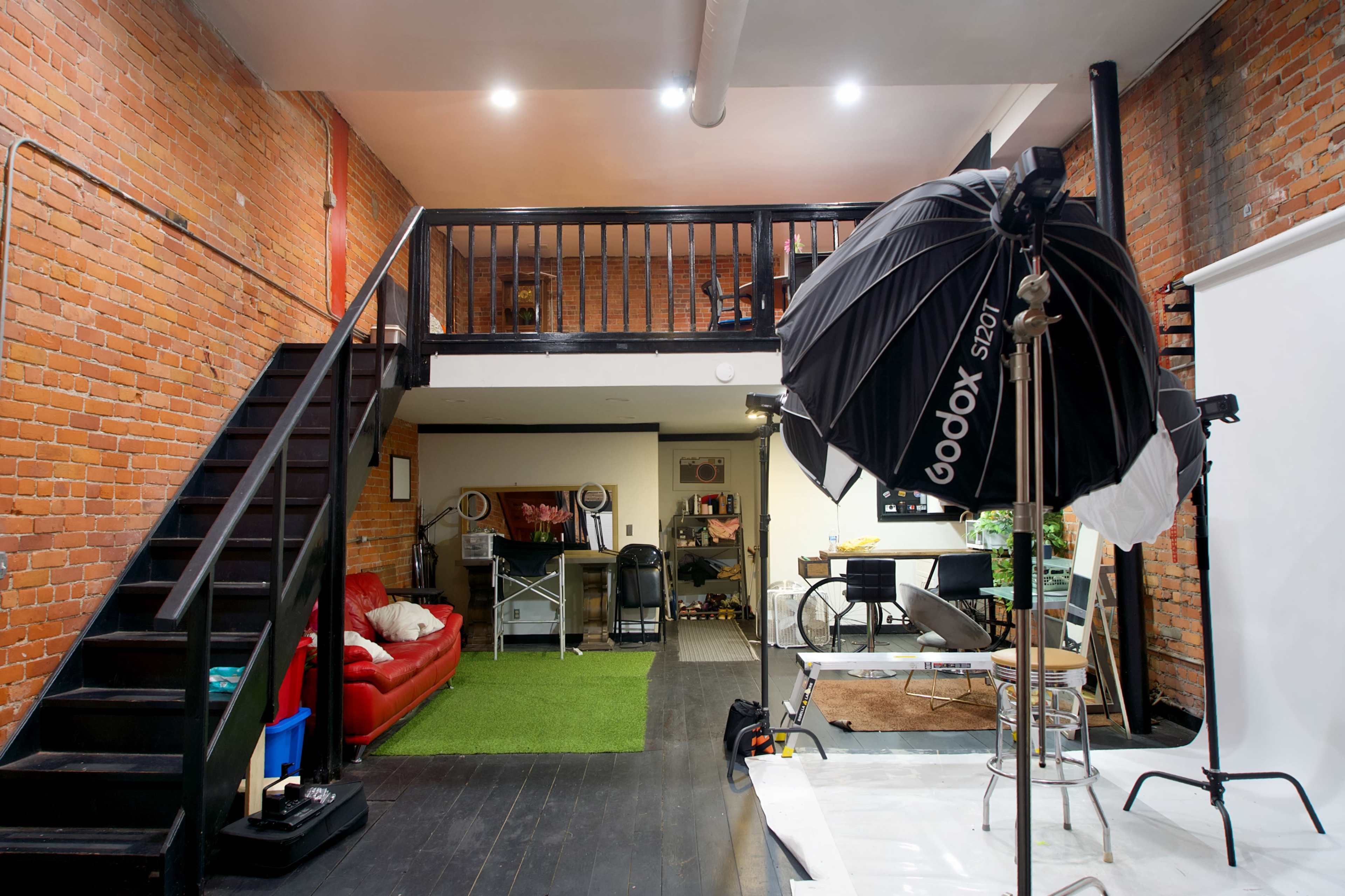 The image shows a spacious photo studio featuring exposed brick walls, a staircase leading to an upper level, and various furniture pieces, including a red sofa and a photography lighting setup.