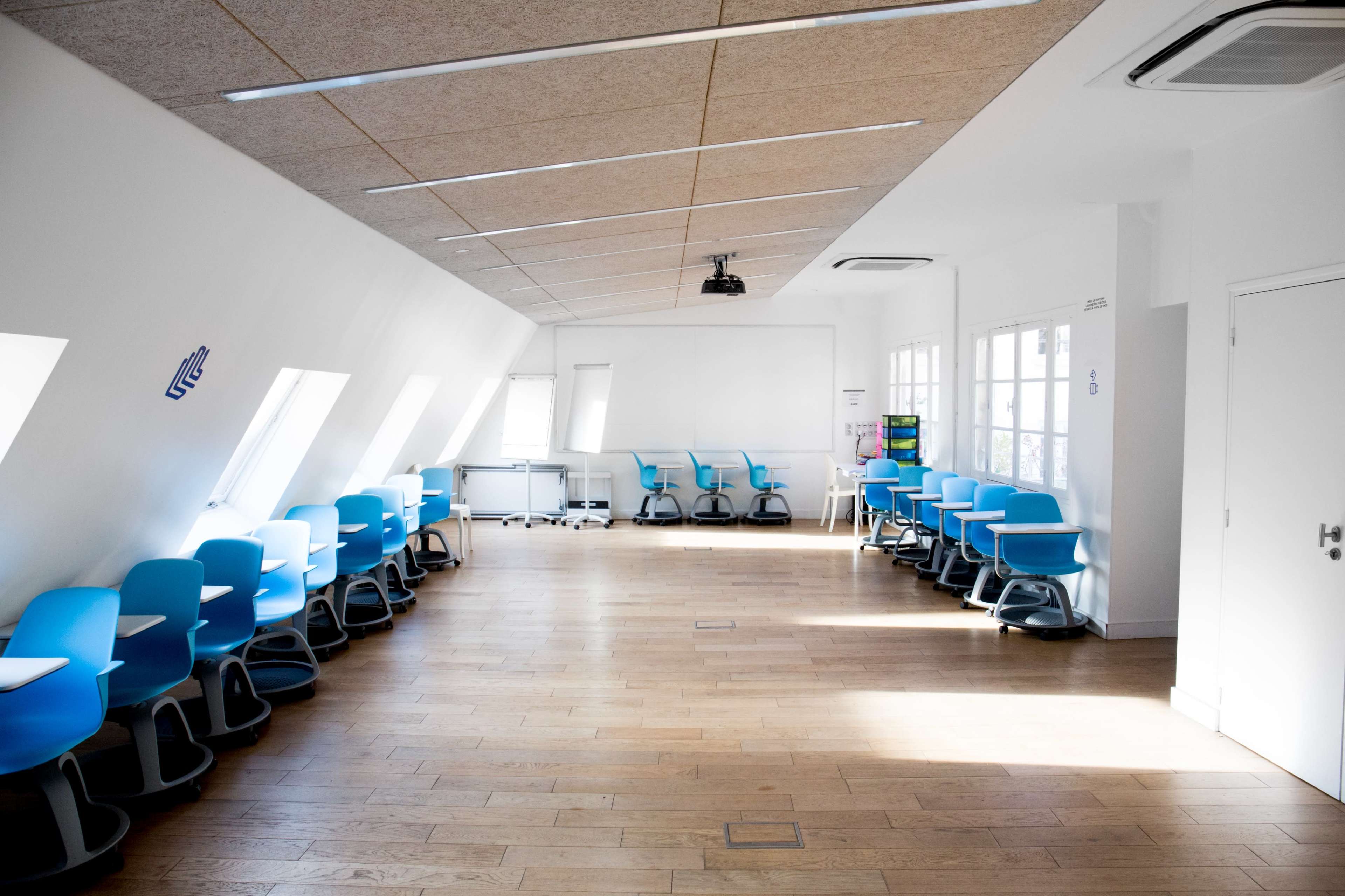 A classroom with blue chairs arranged in rows along the walls and wooden flooring, illuminated by natural light from windows.