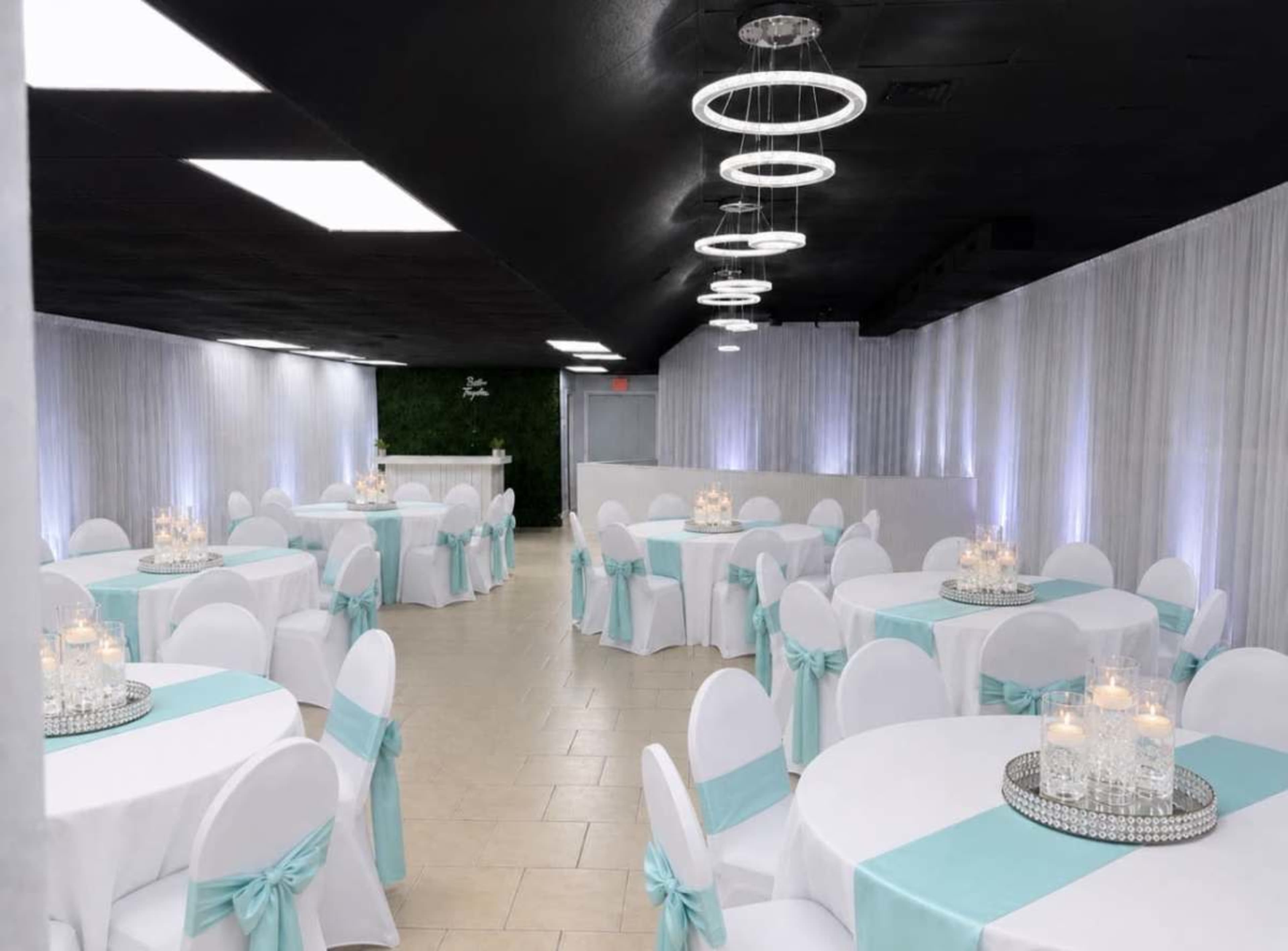 The image shows a banquet hall arranged with round tables covered in white tablecloths and accented with turquoise sashes on the chairs, illuminated by soft lighting.