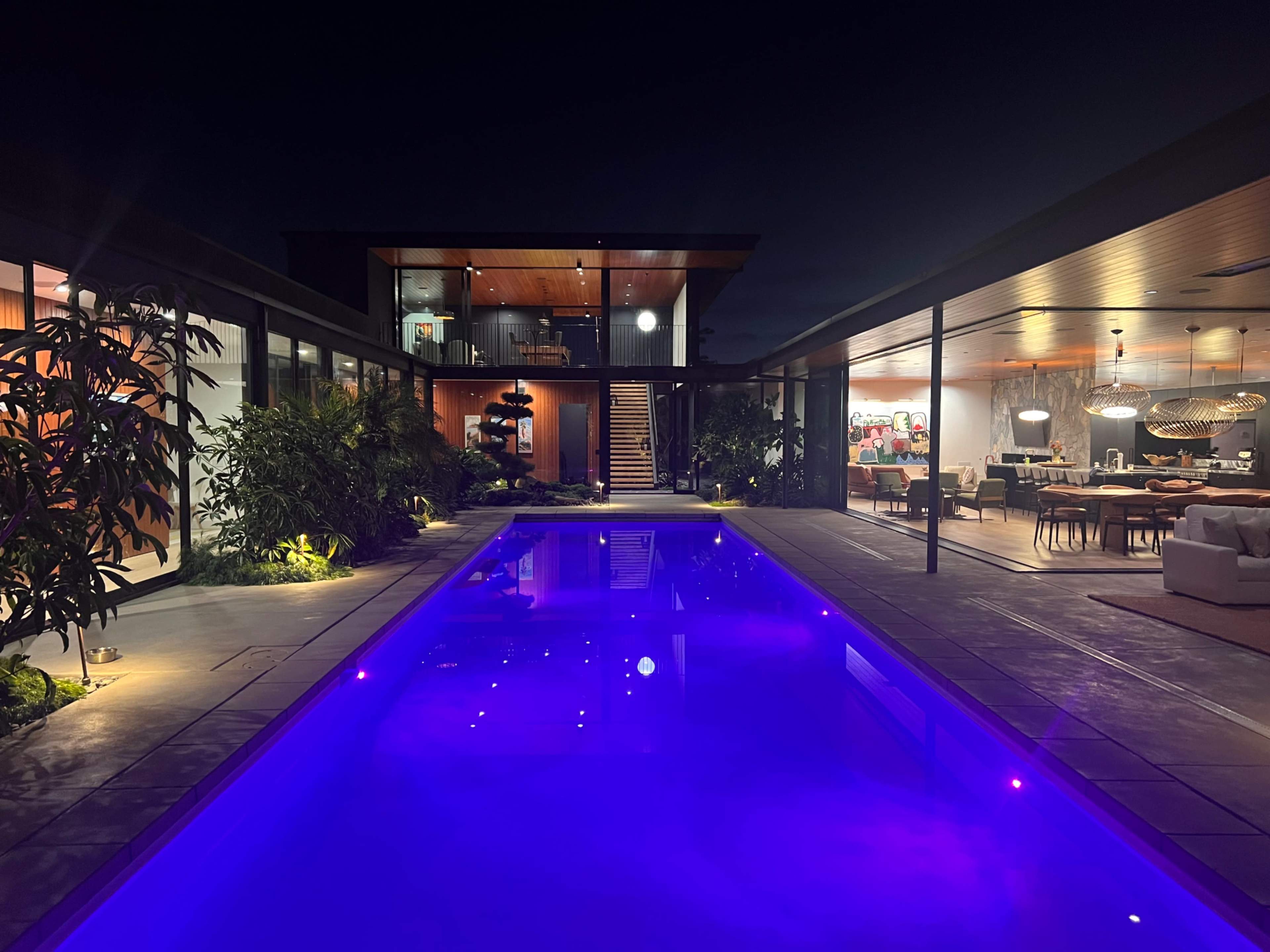 A modern house with a sleek design features a violet-lit pool surrounded by tropical plants, with an open living area visible through large windows.