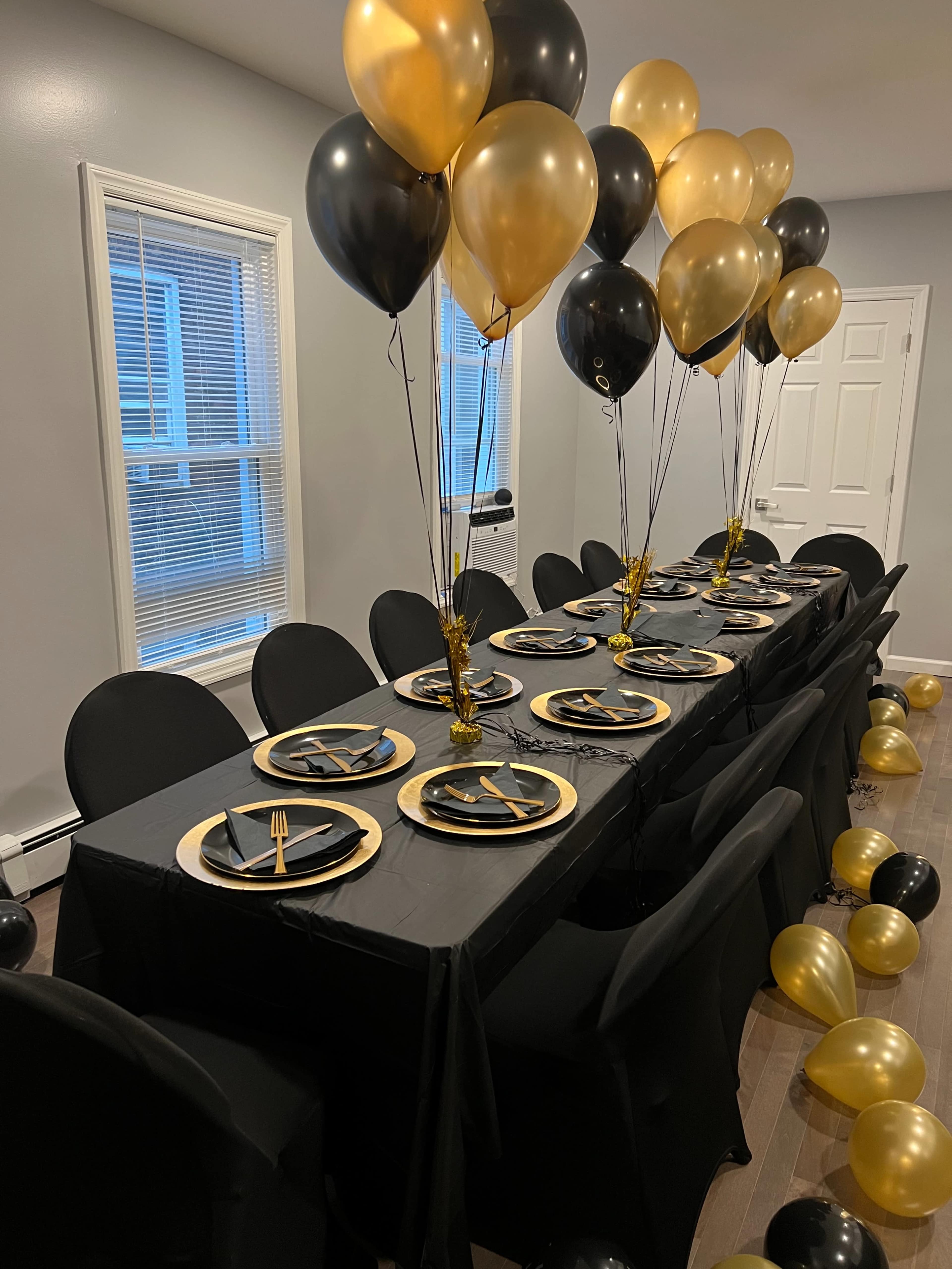 A long dining table is set with black and gold tableware, surrounded by black chairs and adorned with black and gold balloons.