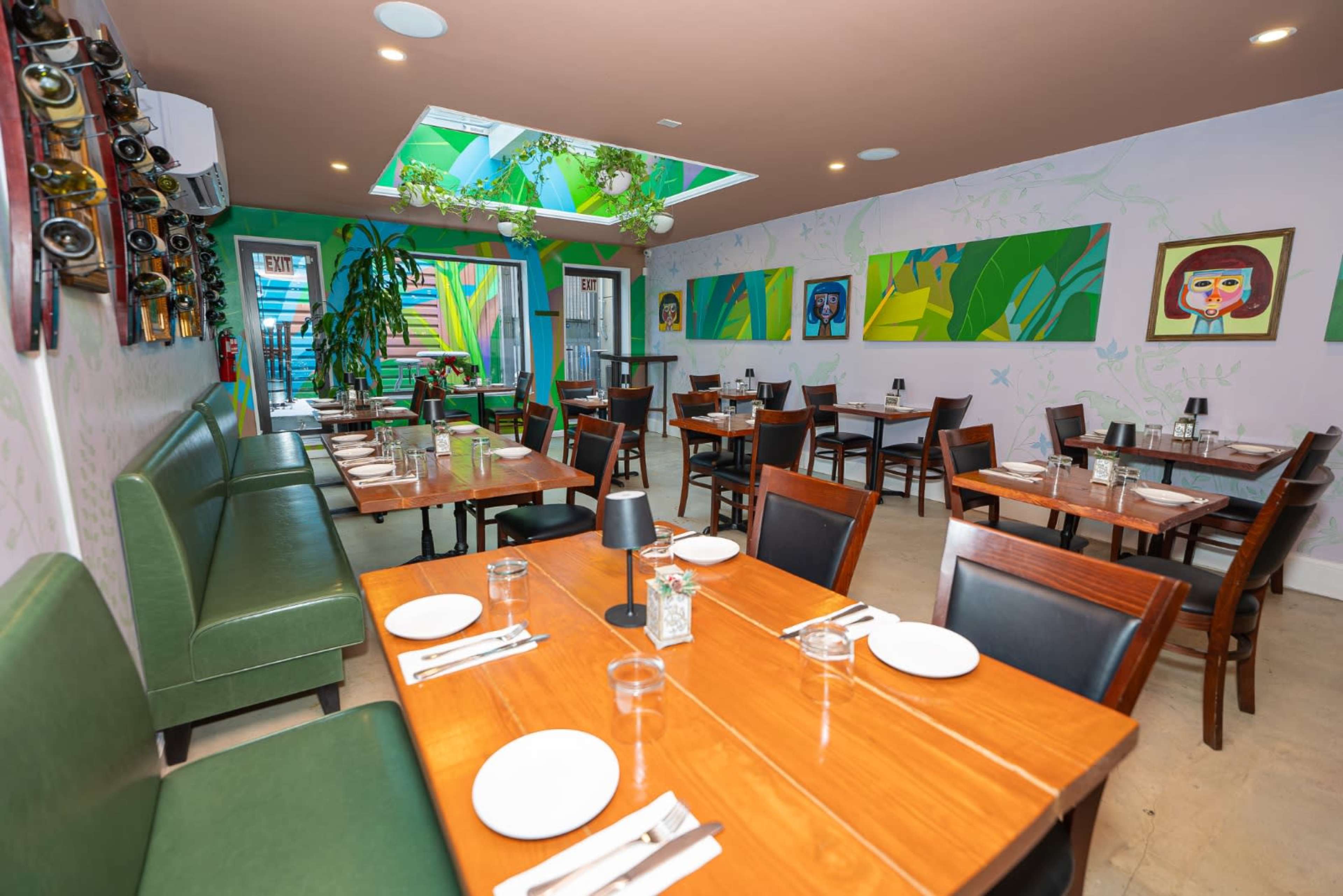 The image shows the interior of a restaurant featuring wooden tables, green seating, colorful wall art, and bright green plants.