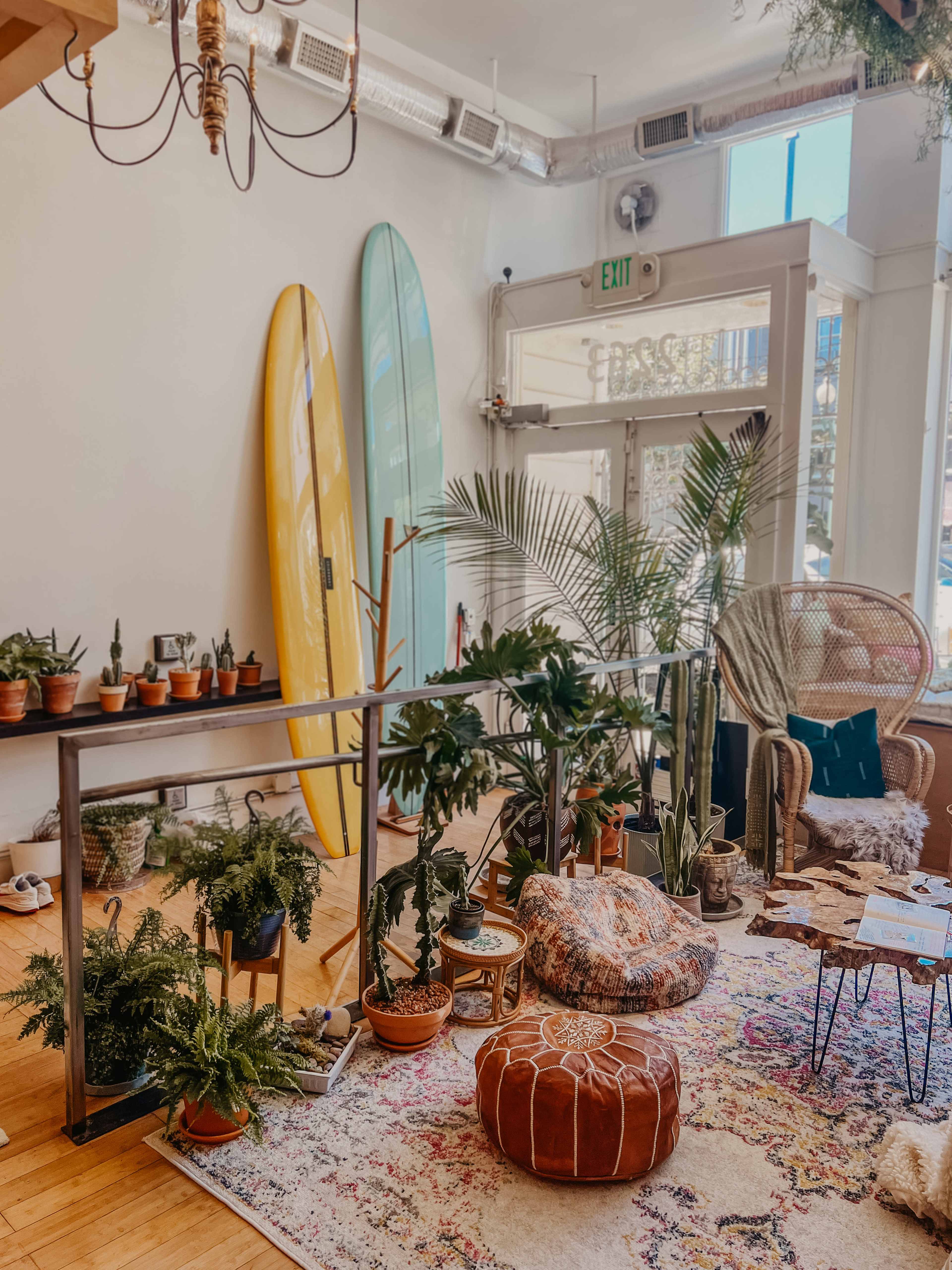 A bright interior with two surfboards leaning against the wall, various potted plants arranged on shelves, and a cozy seating area featuring a woven chair and a decorative pouf.