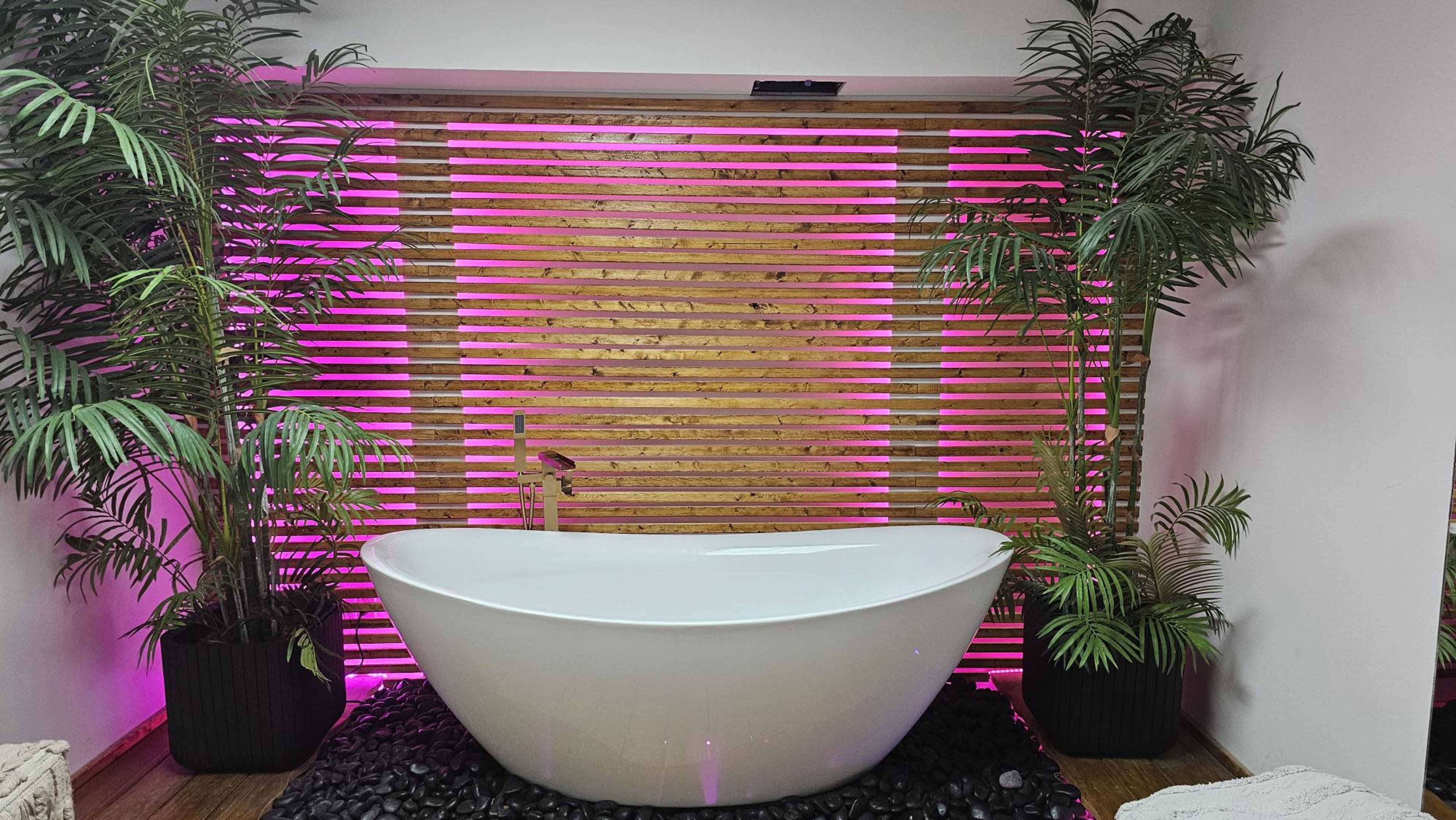 The image features a white freestanding bathtub positioned in front of a wooden slatted wall illuminated with pink light, surrounded by potted plants.
