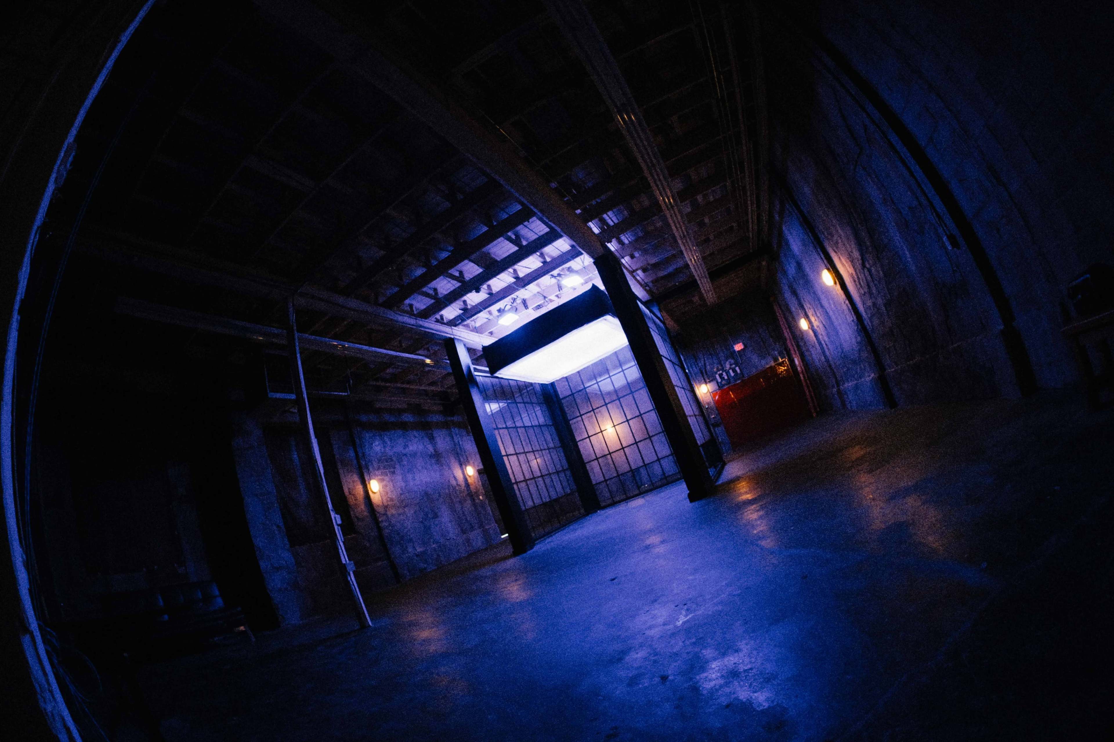 The image shows a dimly lit industrial space with high ceilings, featuring a large window at the far end that lets in a blue light.