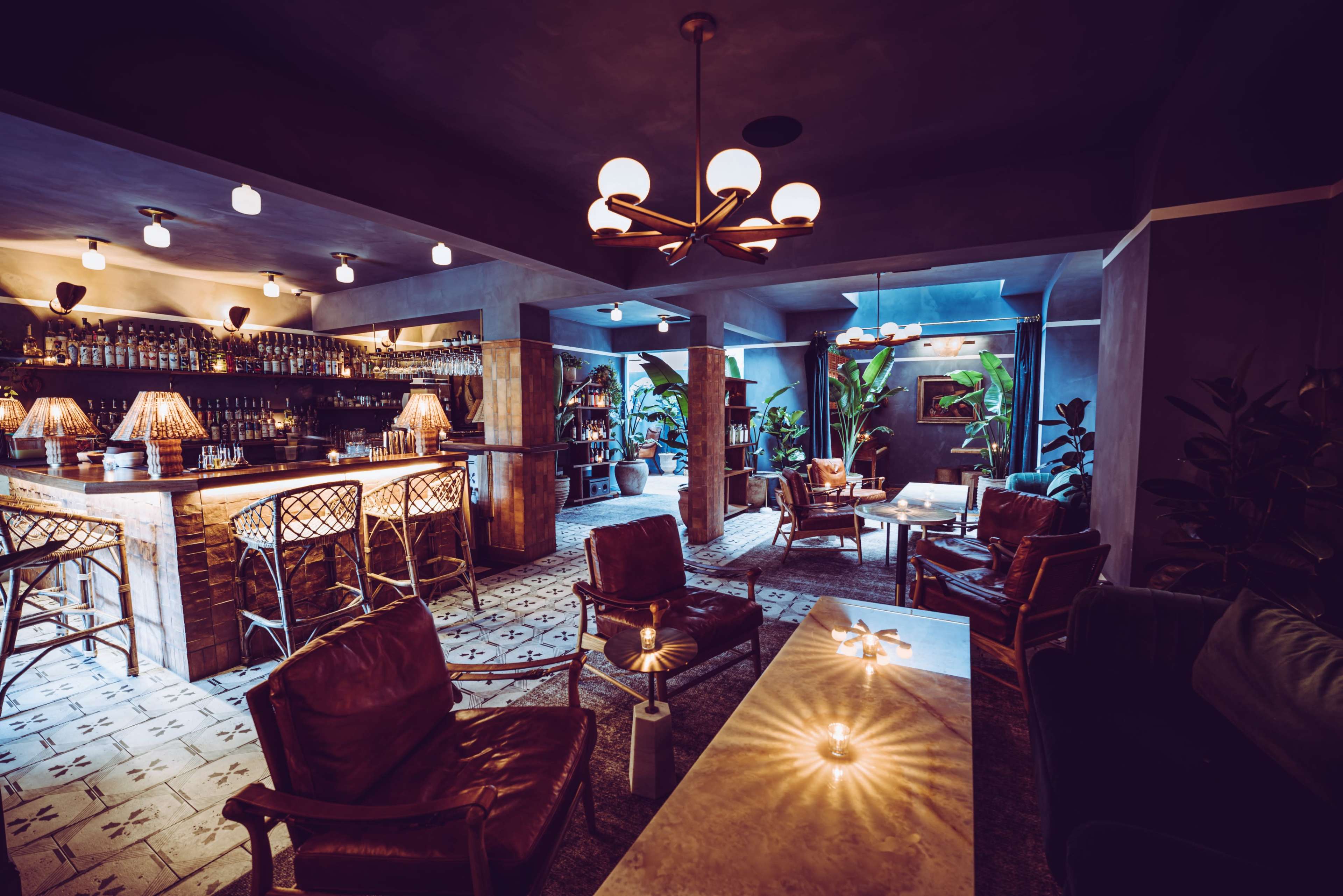 The image depicts a stylish bar interior with a marble table, leather seating, a well-stocked counter, and dim lighting accentuated by plants and decorative lamps.