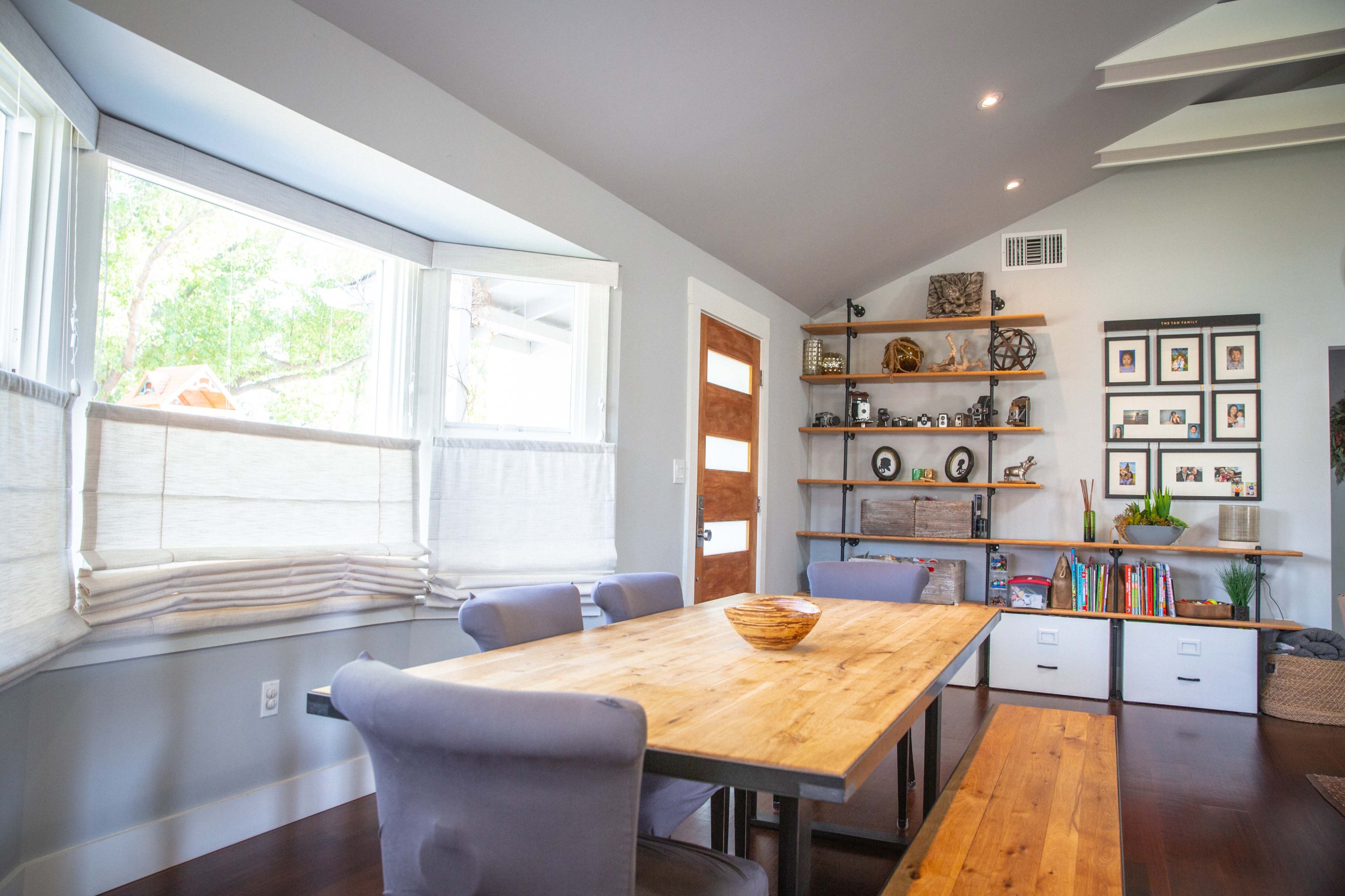 A dining area with a wooden table surrounded by chairs, large windows allowing natural light, and a wall shelf displaying various items and framed pictures.