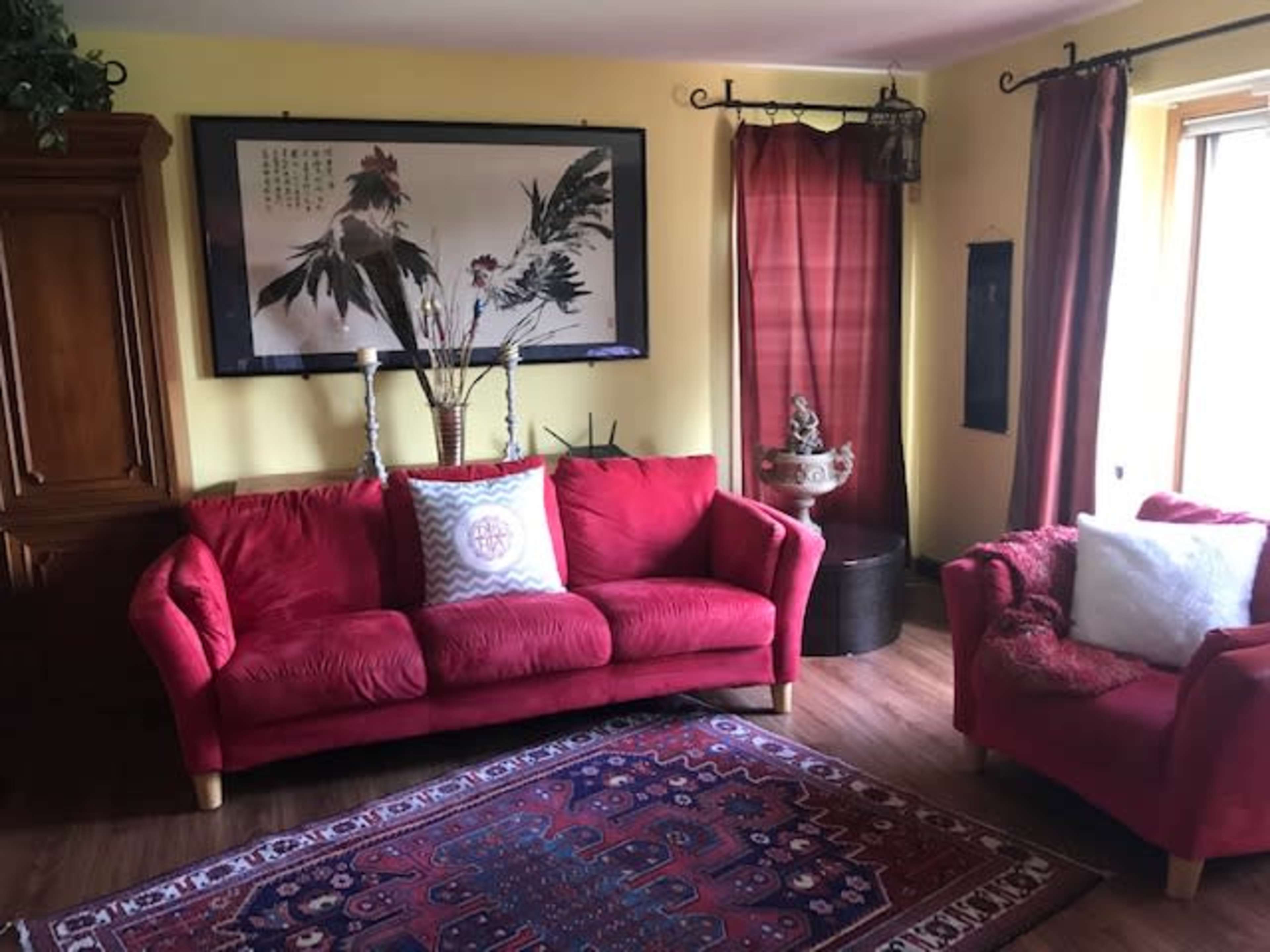 The image shows a living room with a red sofa, a small red armchair, a decorative rug, and a large framed artwork on the wall.