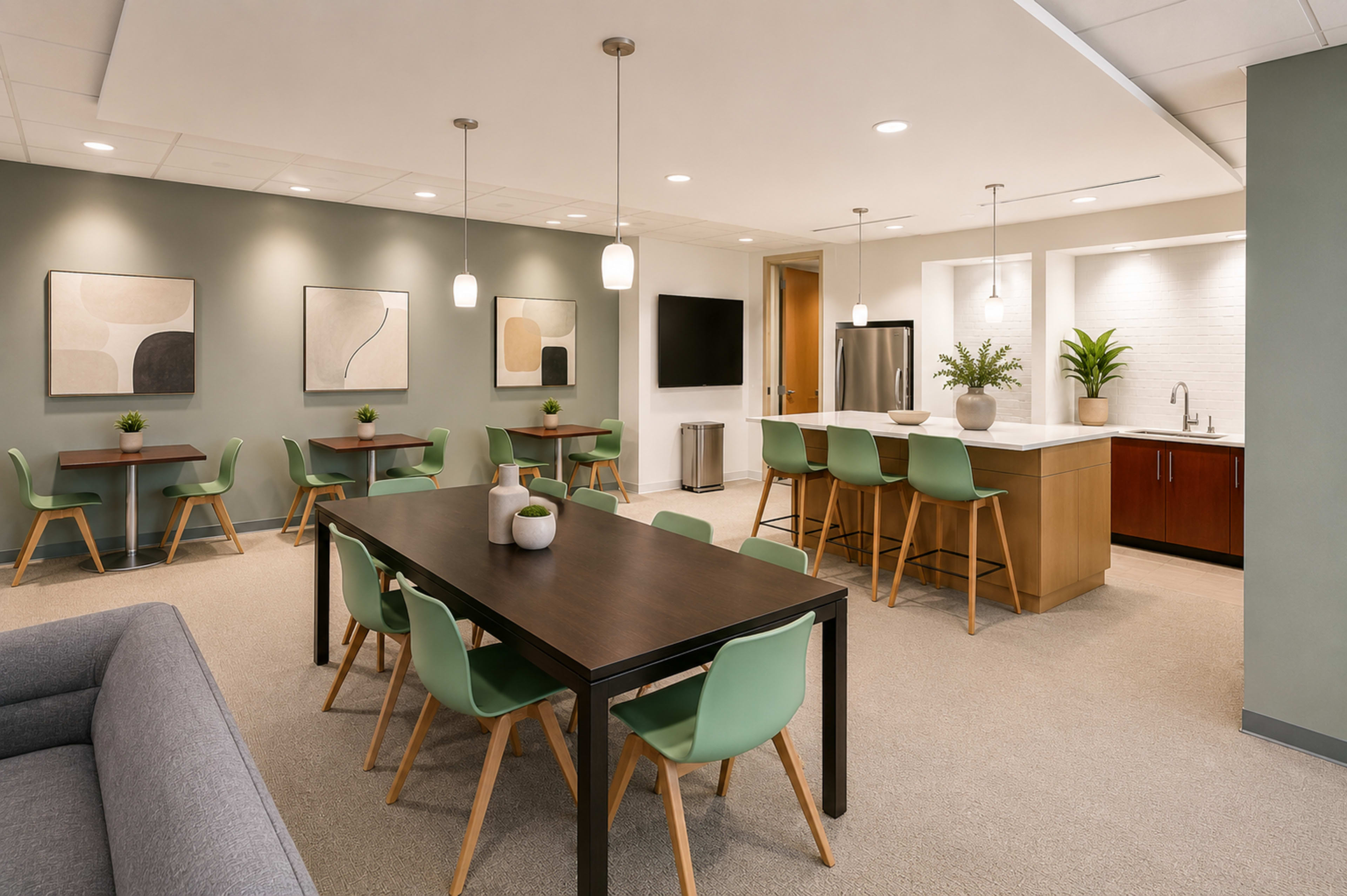 The image shows a modern break room with a seating area featuring tables, chairs, a kitchenette, and decorative plants.