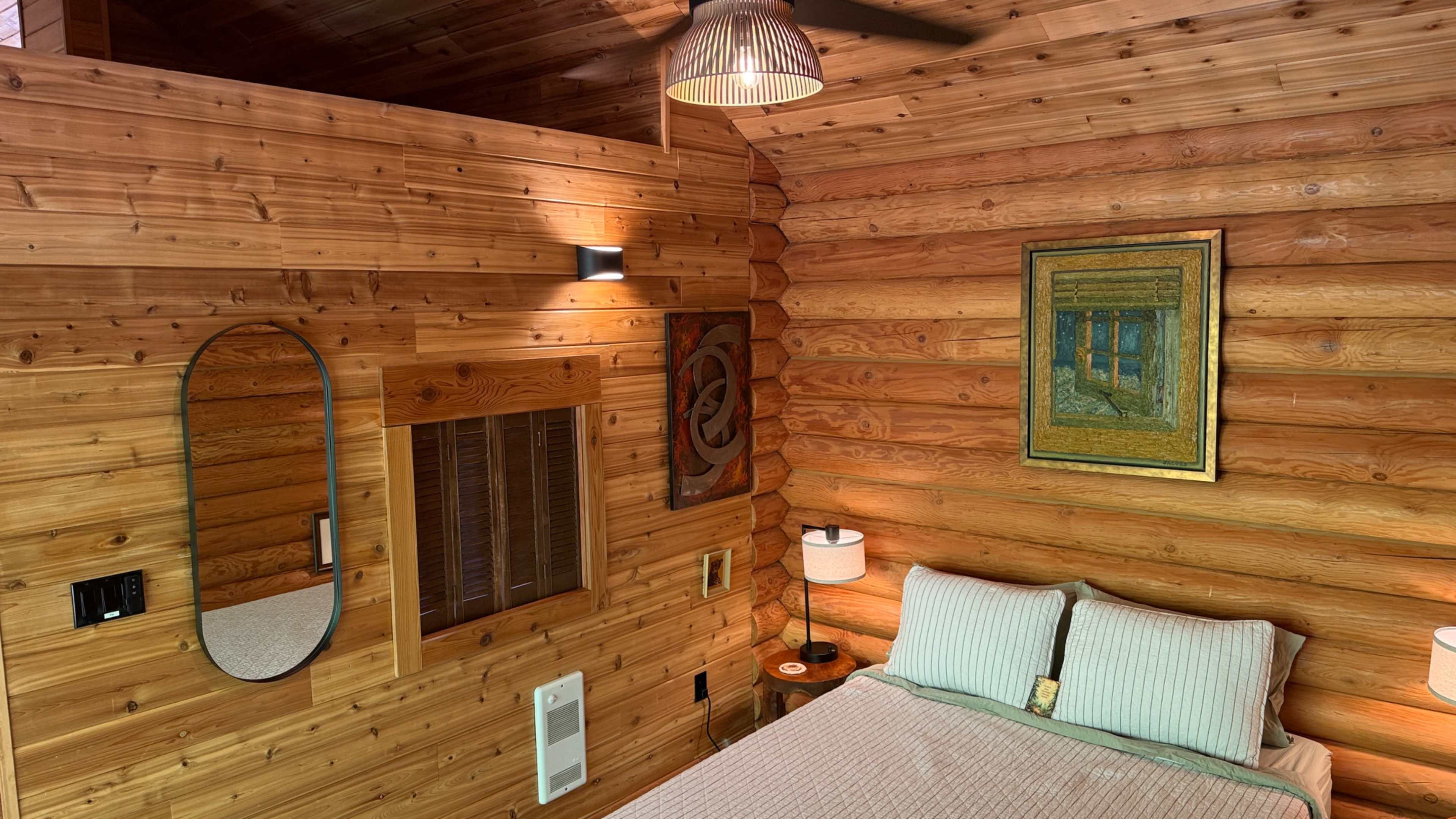The image shows a cozy wooden cabin bedroom featuring a wooden wall, a mirror, a painting, bedside lamps, and a neatly made bed.