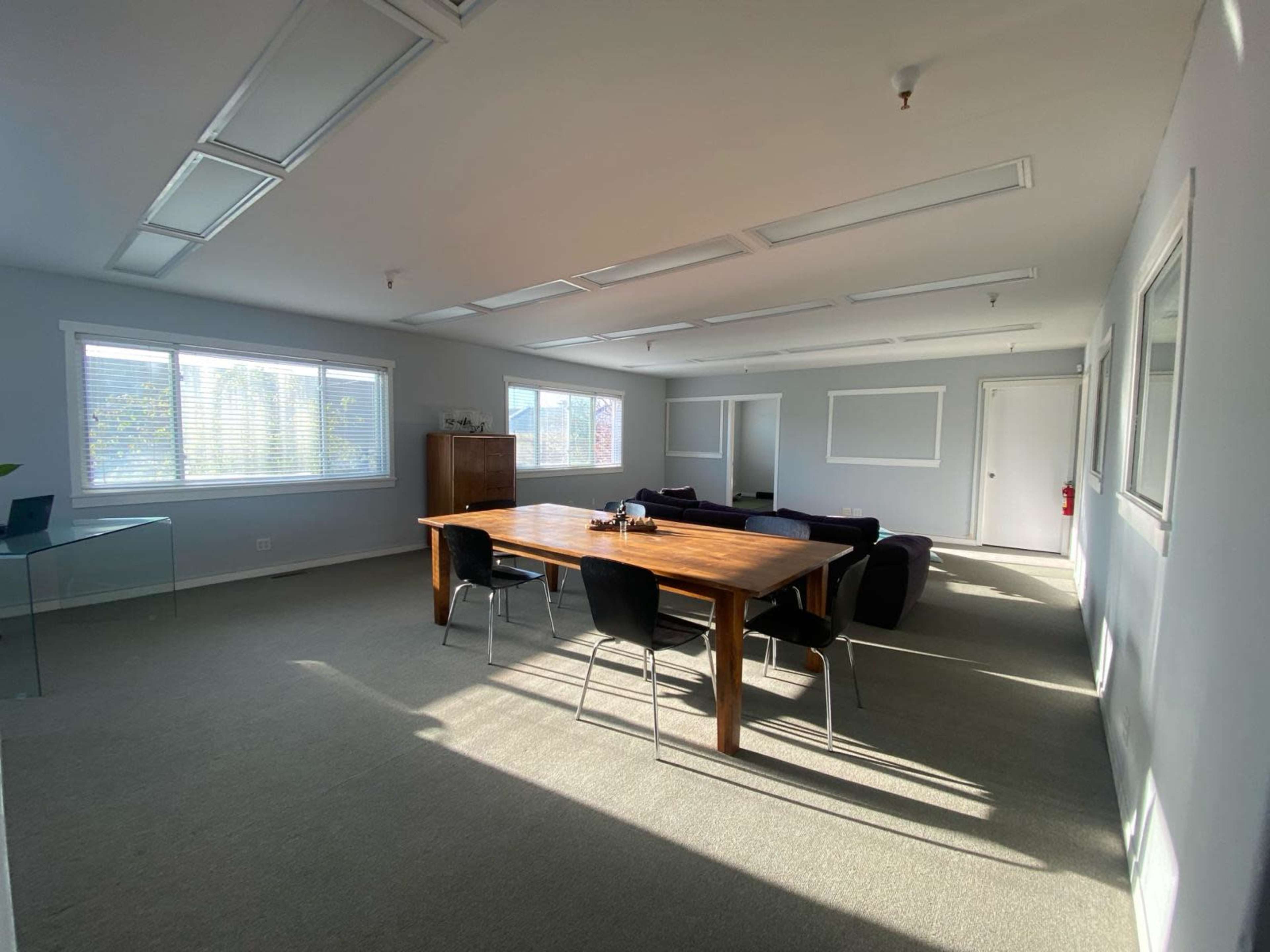 A spacious office features a wooden table surrounded by chairs, with large windows illuminating the carpeted floor.