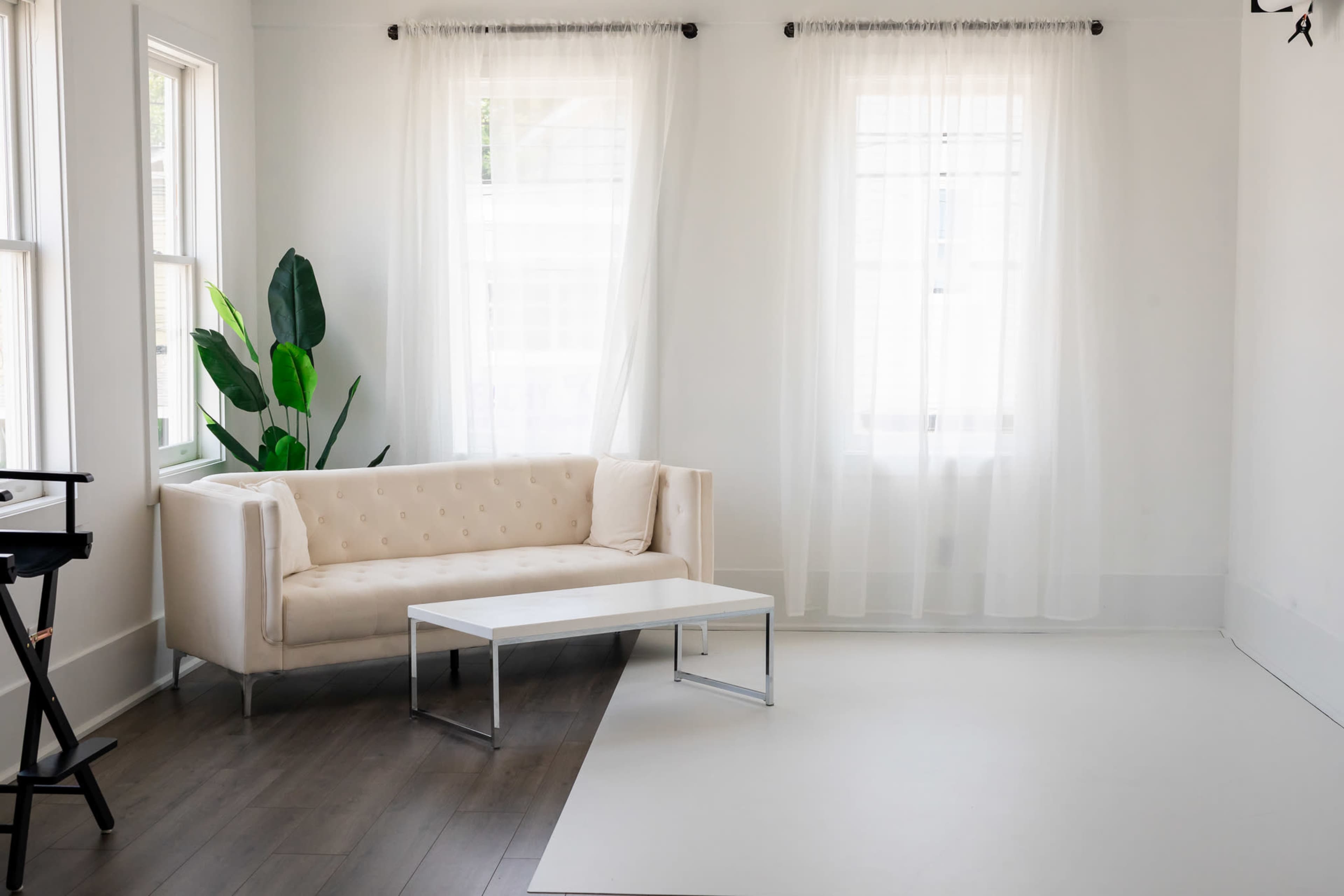 The image shows a minimalist, well-lit room featuring a beige tufted sofa, a small white coffee table, and a large green plant near a window with sheer curtains.