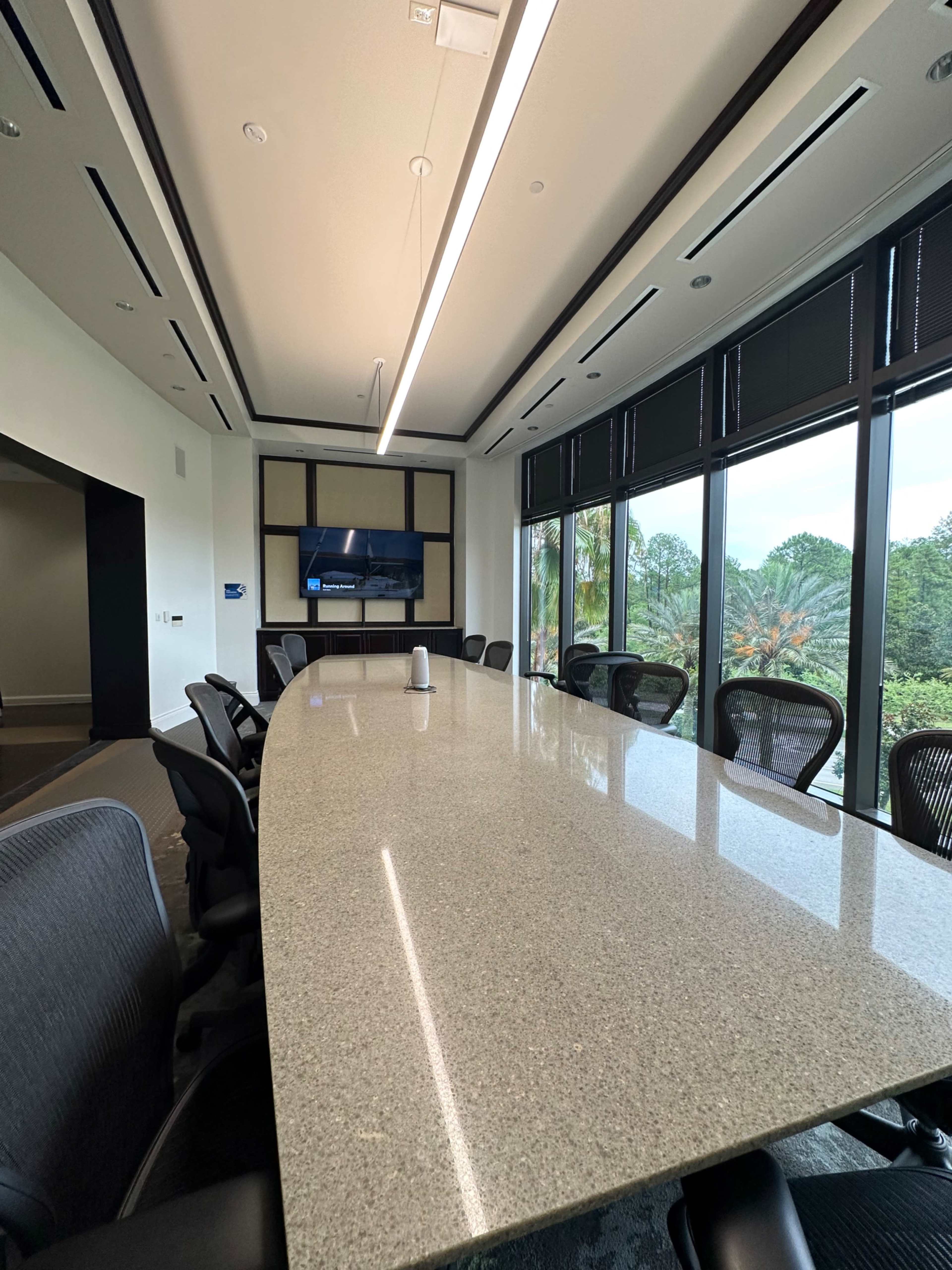 A modern conference room features a long granite table, ergonomic chairs, and large windows overlooking a green landscape.