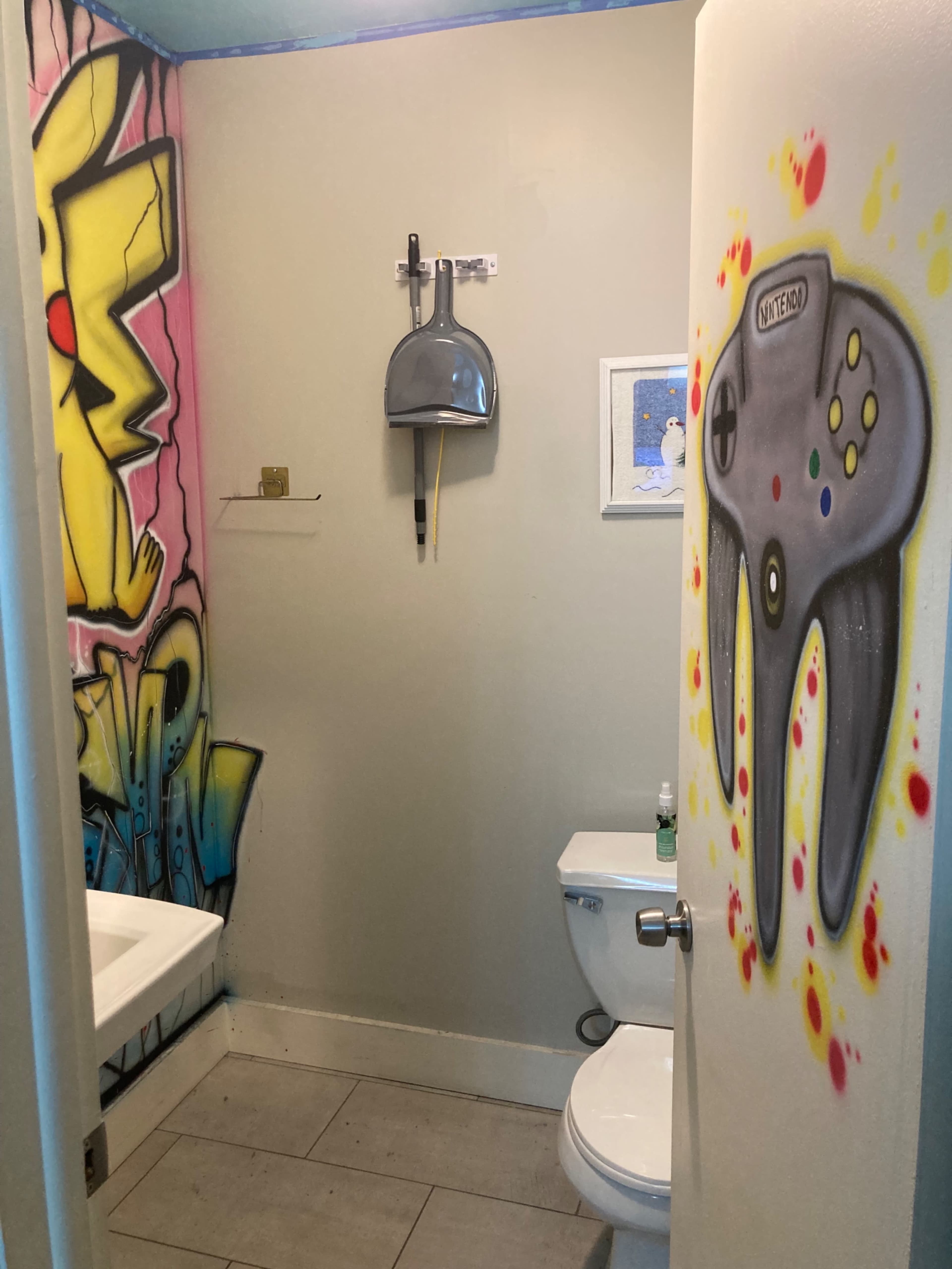 The image shows a small bathroom with graffiti artwork on the walls, including a Pikachu and a Nintendo game controller design.