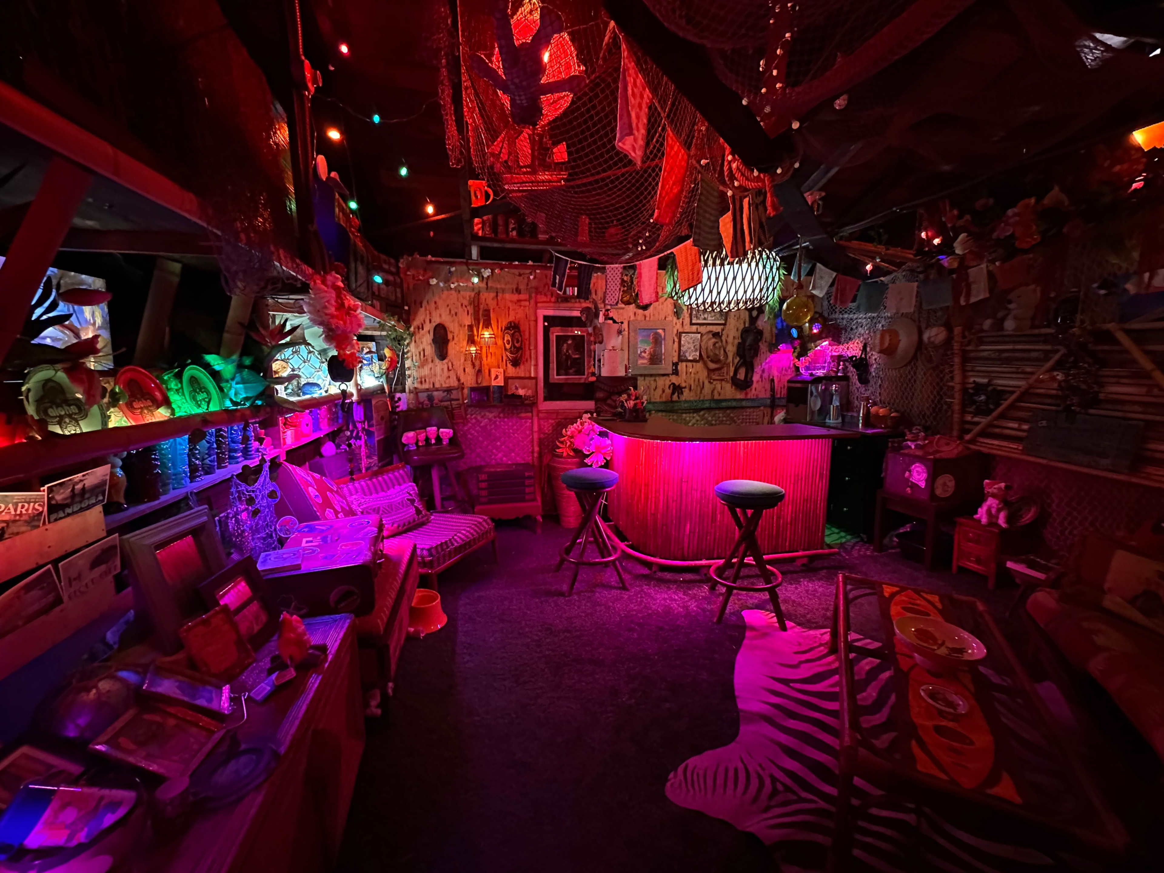 A dimly lit, eclectic bar adorned with colorful lights, decorative nets, and various framed pictures, creating a vibrant and inviting atmosphere.