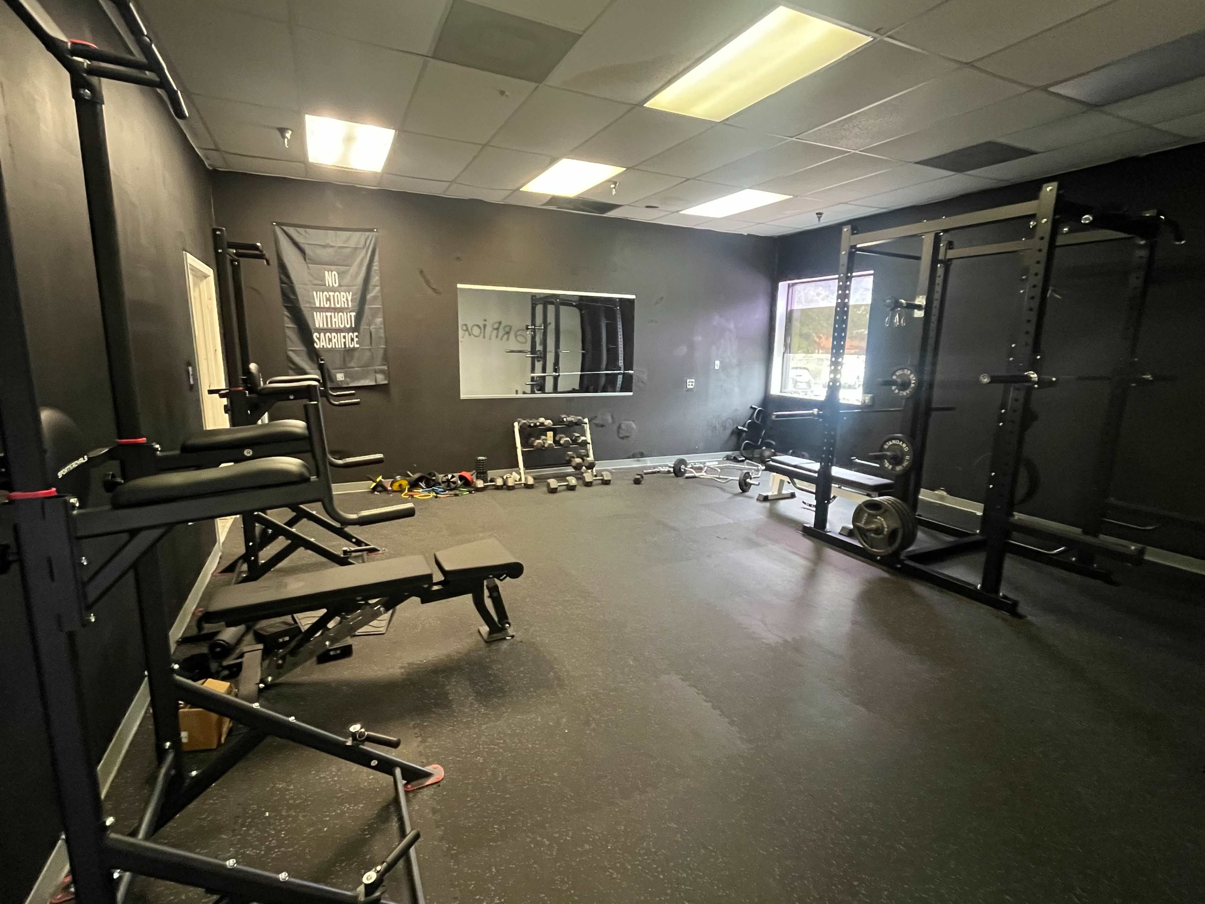 The image shows a gym interior with a weight bench, a squat rack, and scattered dumbbells on the floor against a dark wall.