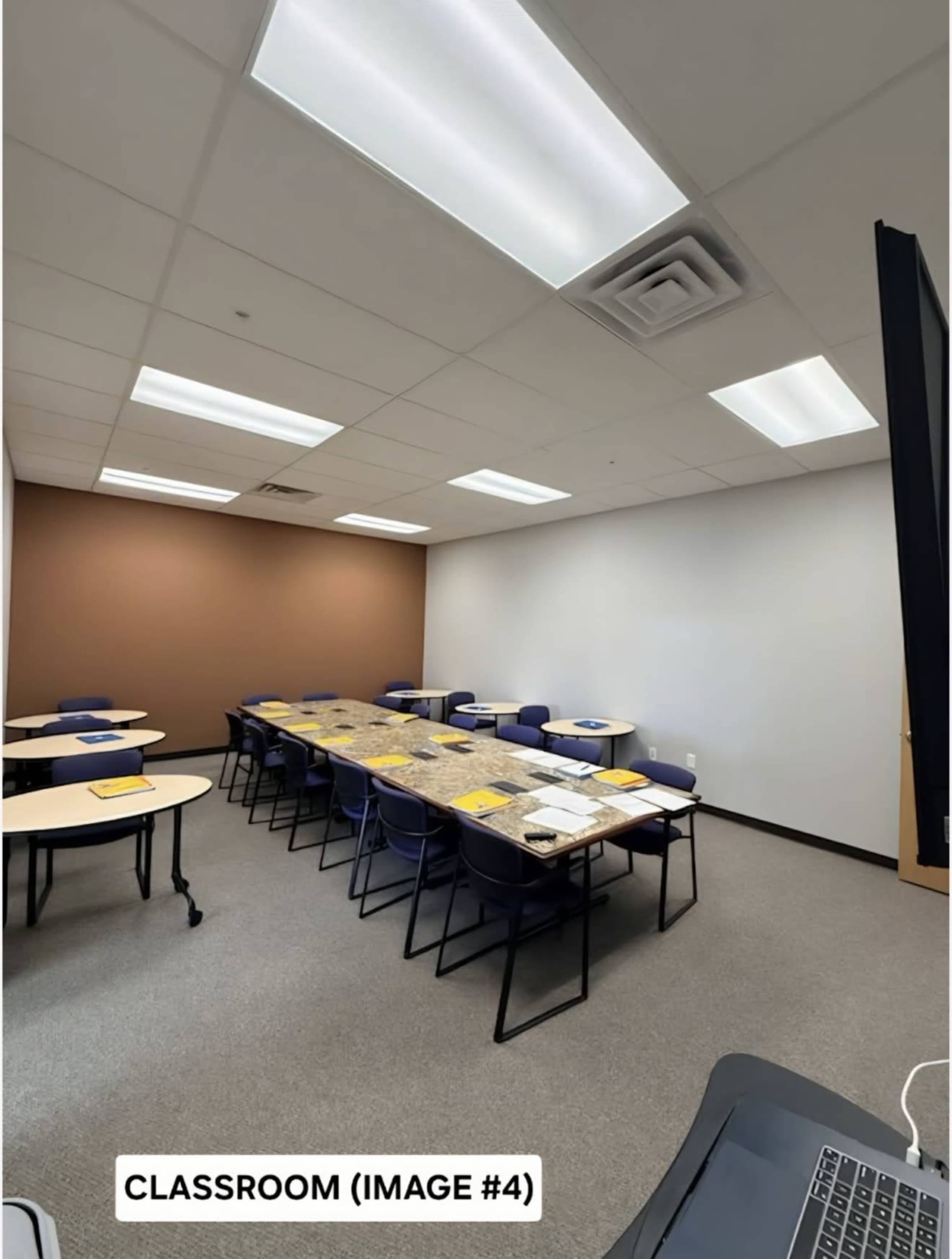 The image shows a classroom with several tables arranged in a U-shape, surrounded by chairs, and a projector screen at the front.