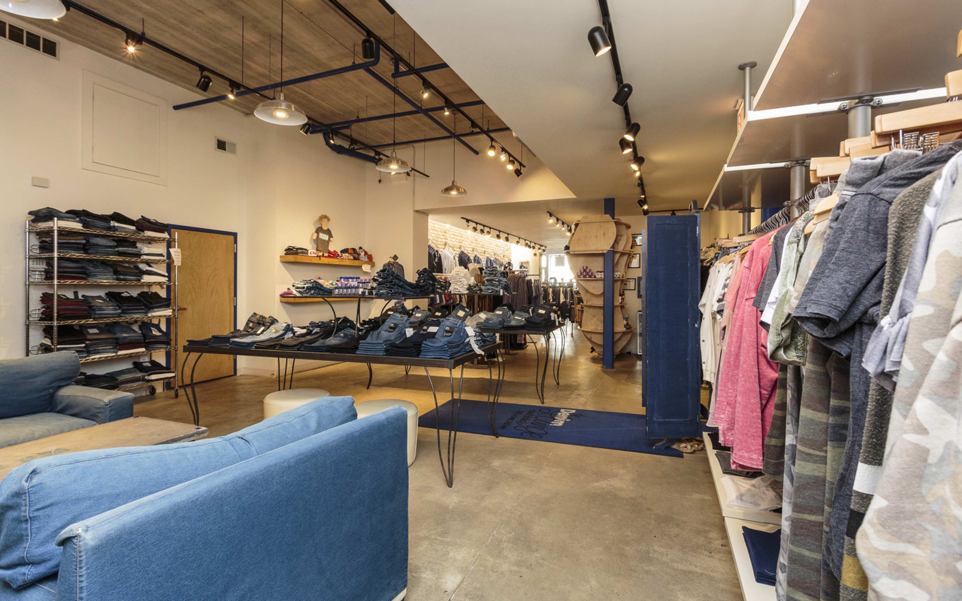 A clothing store displays racks of apparel and organized stacks of jeans on tables, with a seating area in the foreground.