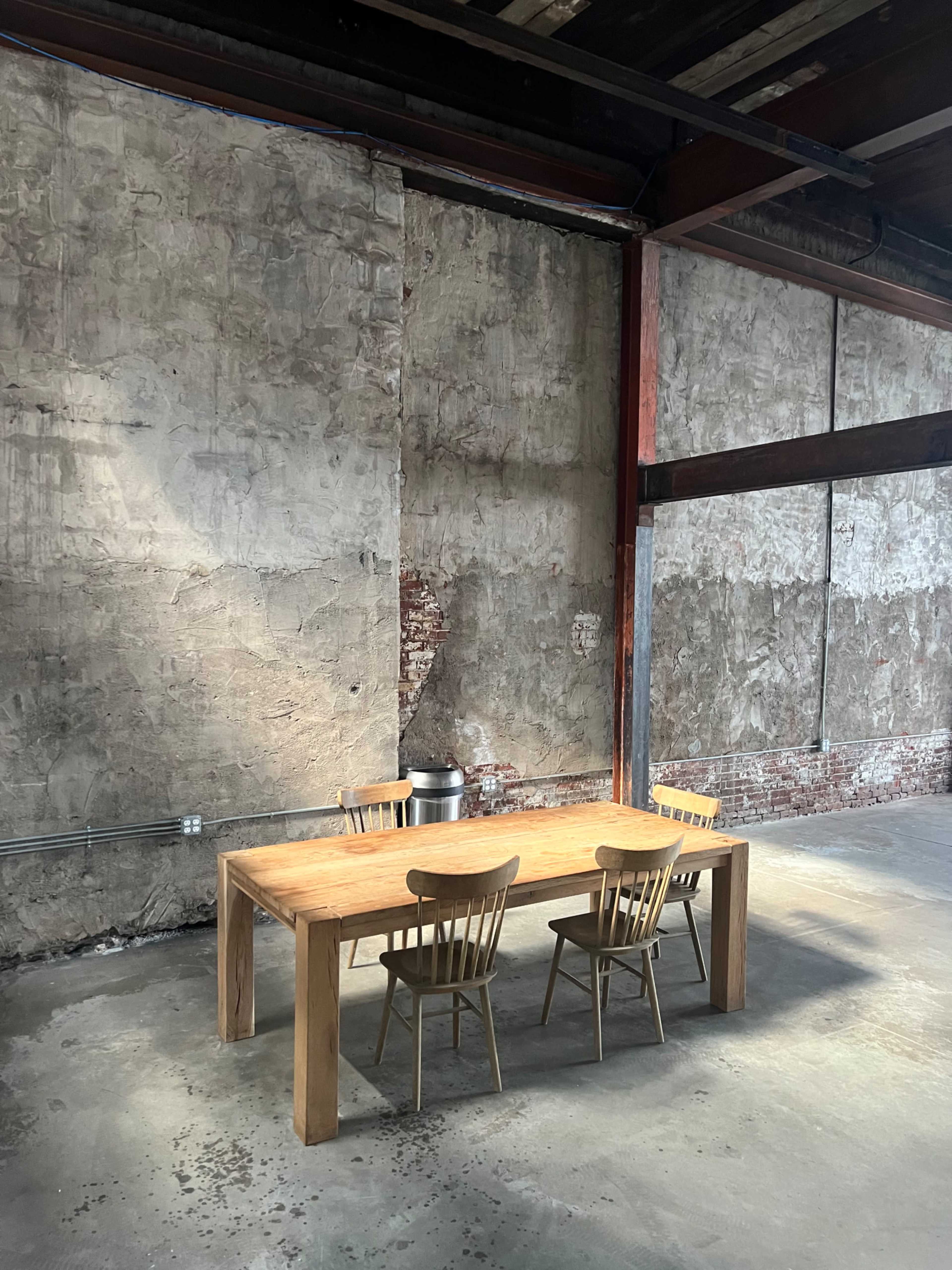 A wooden table with three chairs is set against a bare, textured wall in an industrial space.