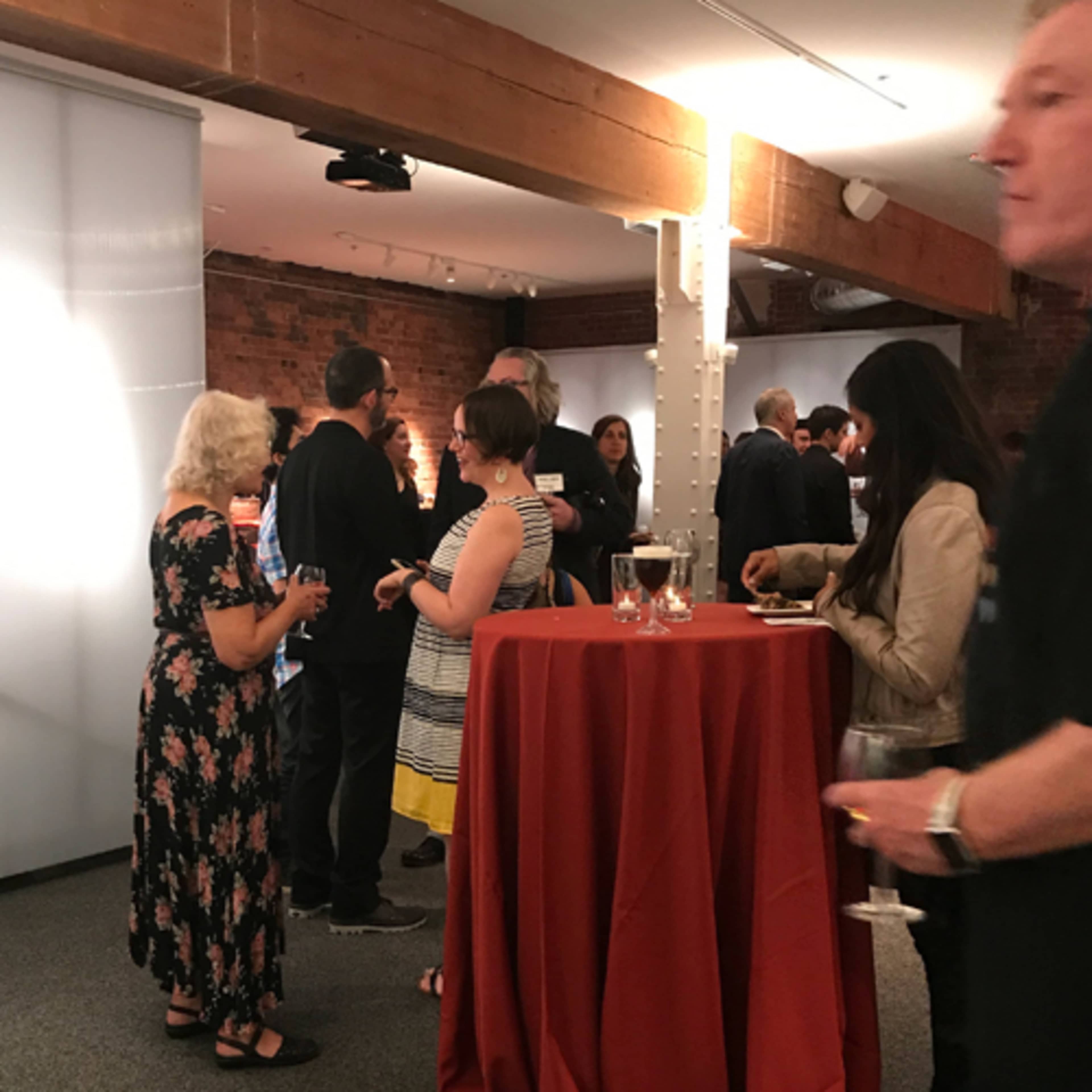 A gathering of people socializing around tall tables in a well-lit indoor space with exposed brick walls and wooden beams.