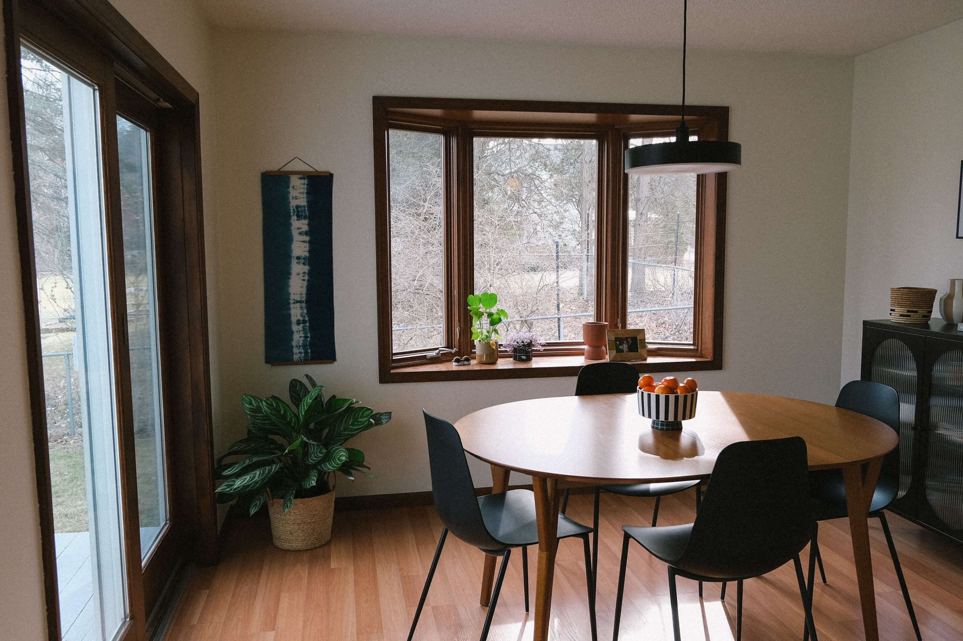 A dining area features a round wooden table surrounded by black chairs set near large windows, with a plant and decorative items on display.